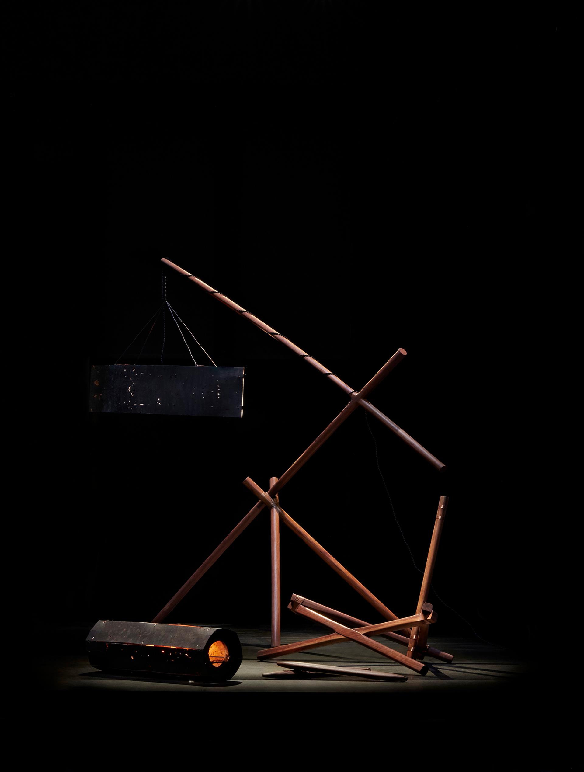 A series of interconnected dark wood beams hold two lights made of black metal. The sculpture is against a dark black background and is spot lit.