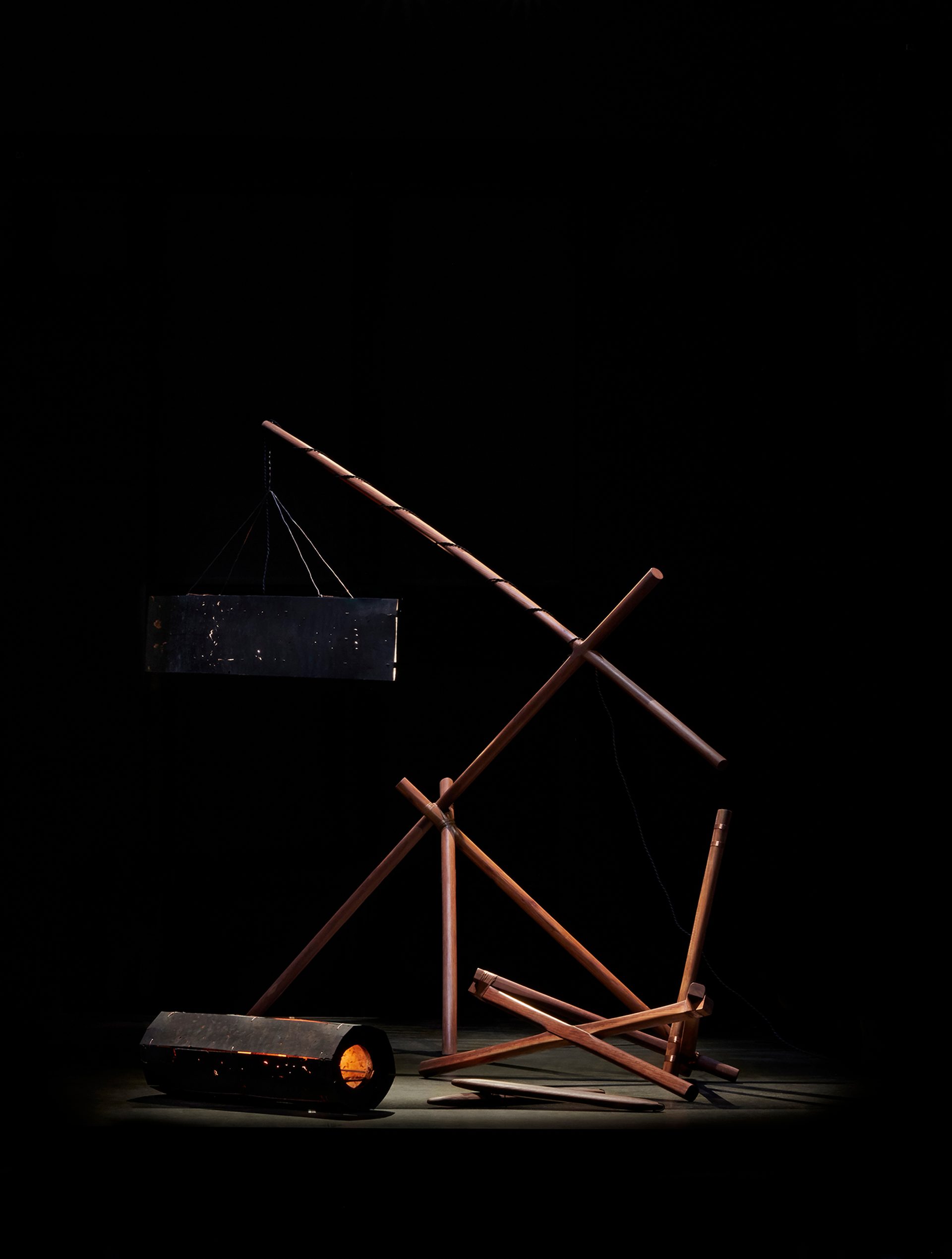 A series of interconnected dark wood beams hold two lights made of black metal. The sculpture is against a dark black background and is spot lit.
