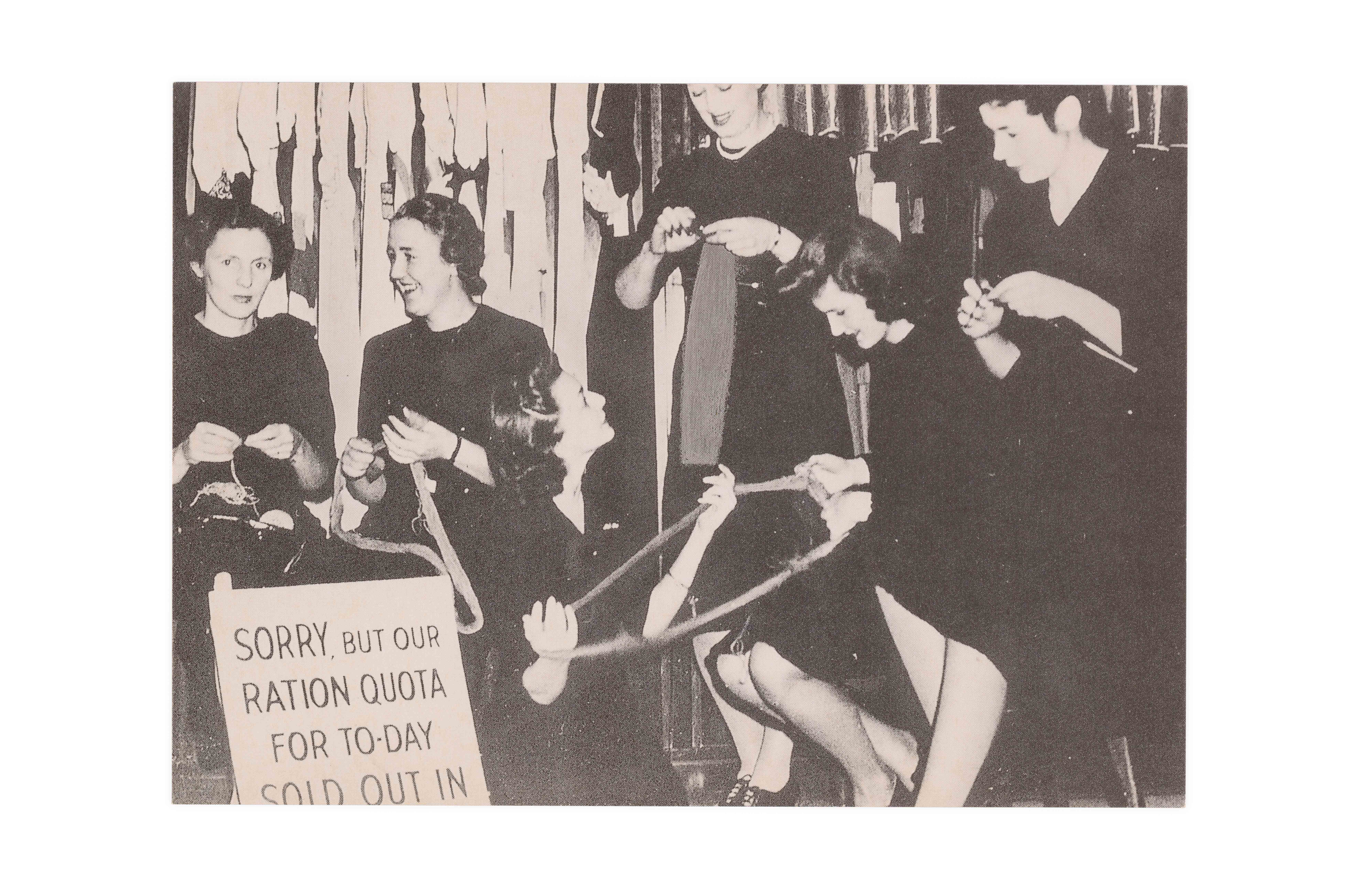 Six women in black work uniforms holding wool and knitting needles. A sign in the front left reads ‘SORRY, BUT OUR RATION QUOTA FOR TO-DAY SOLD OUT IN’