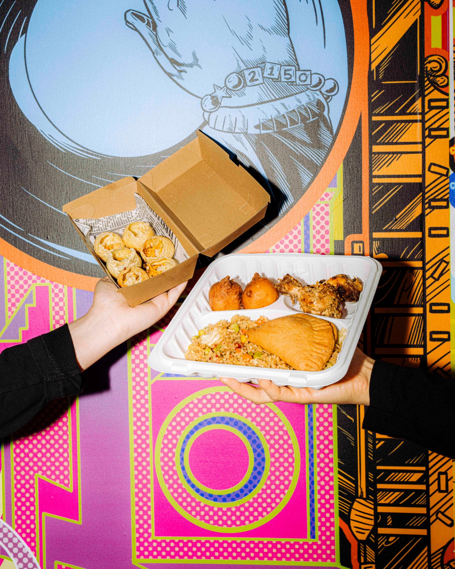 One hand holds up a cardboard box of golden round pieces of food; another lifts a white tray of assorted dishes, all set against a colourful mural backdrop.