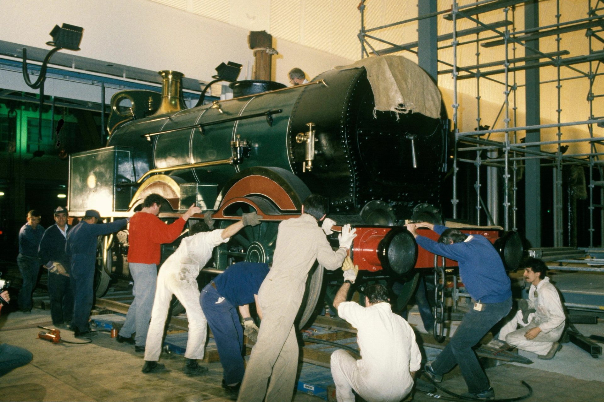 A group of workers surround the front engine of a steam train to pull and push it on a track into the museum.