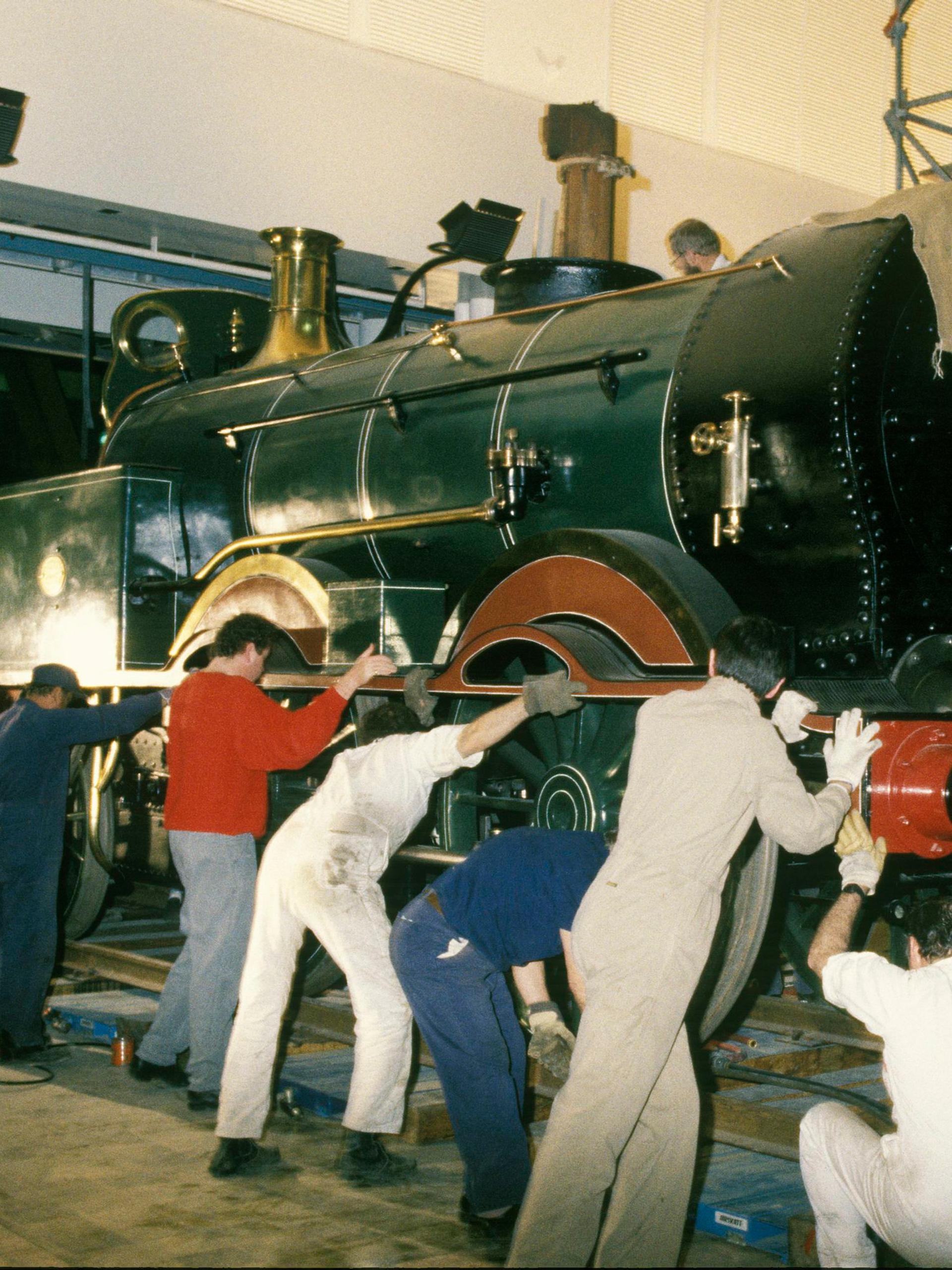 A group of workers surround the front engine of a steam train to pull and push it on a track into the museum.