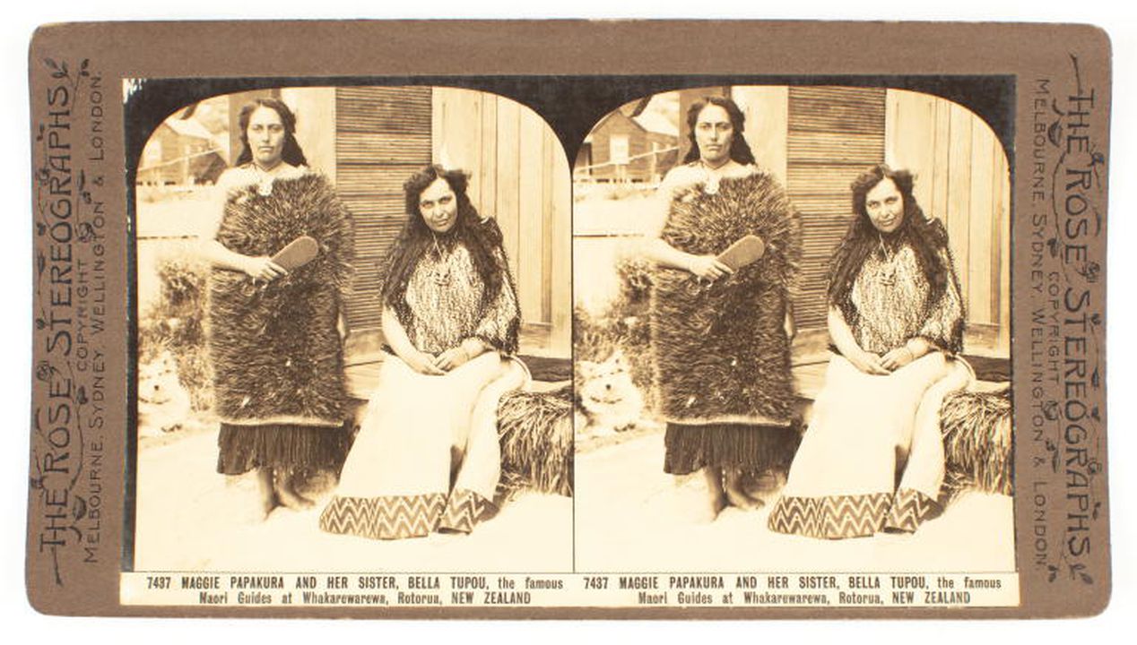 Two Māori women in traditional dress are outside a hut. They have long dark hair. The woman on the right is sitting on the porch or the house. The other woman is standing beside her. She holds a patu. The photograph is sepia.