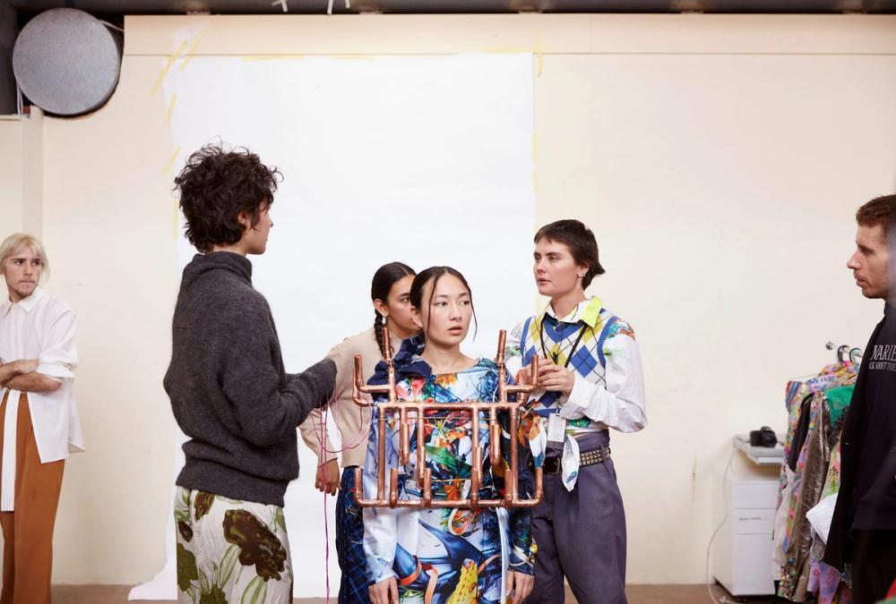 A figure wearing an elaborate sculptural vest made of copper pipes stands amongst a group of people talking to each other.