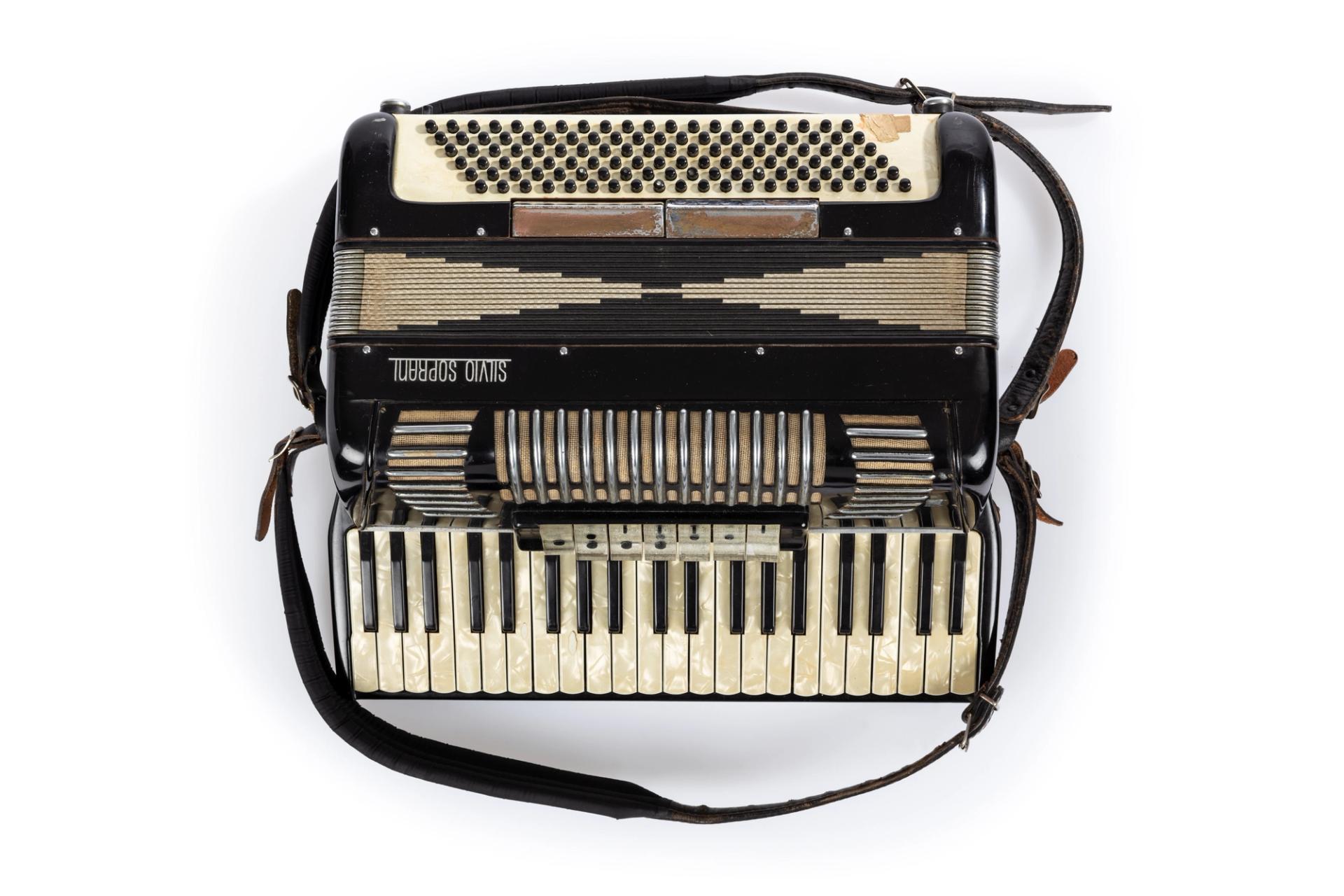 Black piano accordian with the brand name 'Silvio Soprani' on a silver metal badge on front. Piano accordian features twenty four ivory keys, seventeen black keys, seven transparent buttons beside the keyboard and one hundred and twenty small circular shaped buttons on the rear side. Two leather and cloth straps with metal buckles are attached to one end.