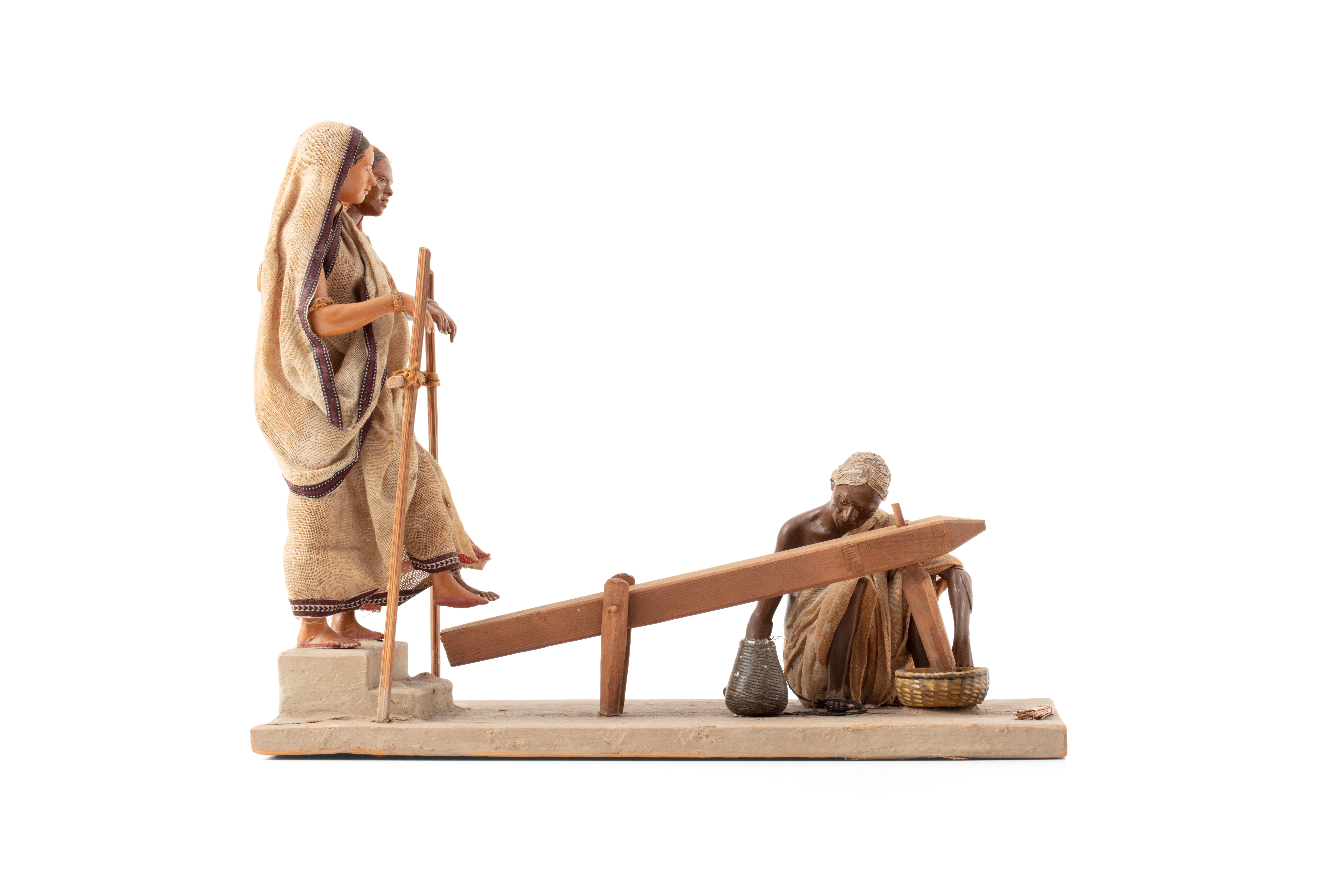 Molded figure of three people grinding rice, made of terracotta. Two women wearing saris stand on one end, using their feet to step on and lift a large wooden panel. At the end of the panel, another figure kneels to a bowl of rice where the panel stabs and grinds the rice.