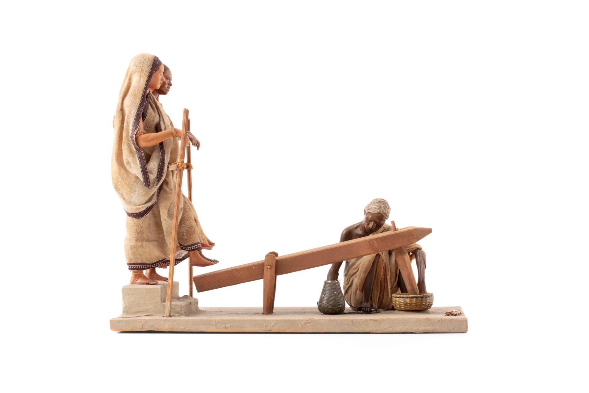 Molded figure of three people grinding rice, made of terracotta. Two women wearing saris stand on one end, using their feet to step on and lift a large wooden panel. At the end of the panel, another figure kneels to a bowl of rice where the panel stabs and grinds the rice.