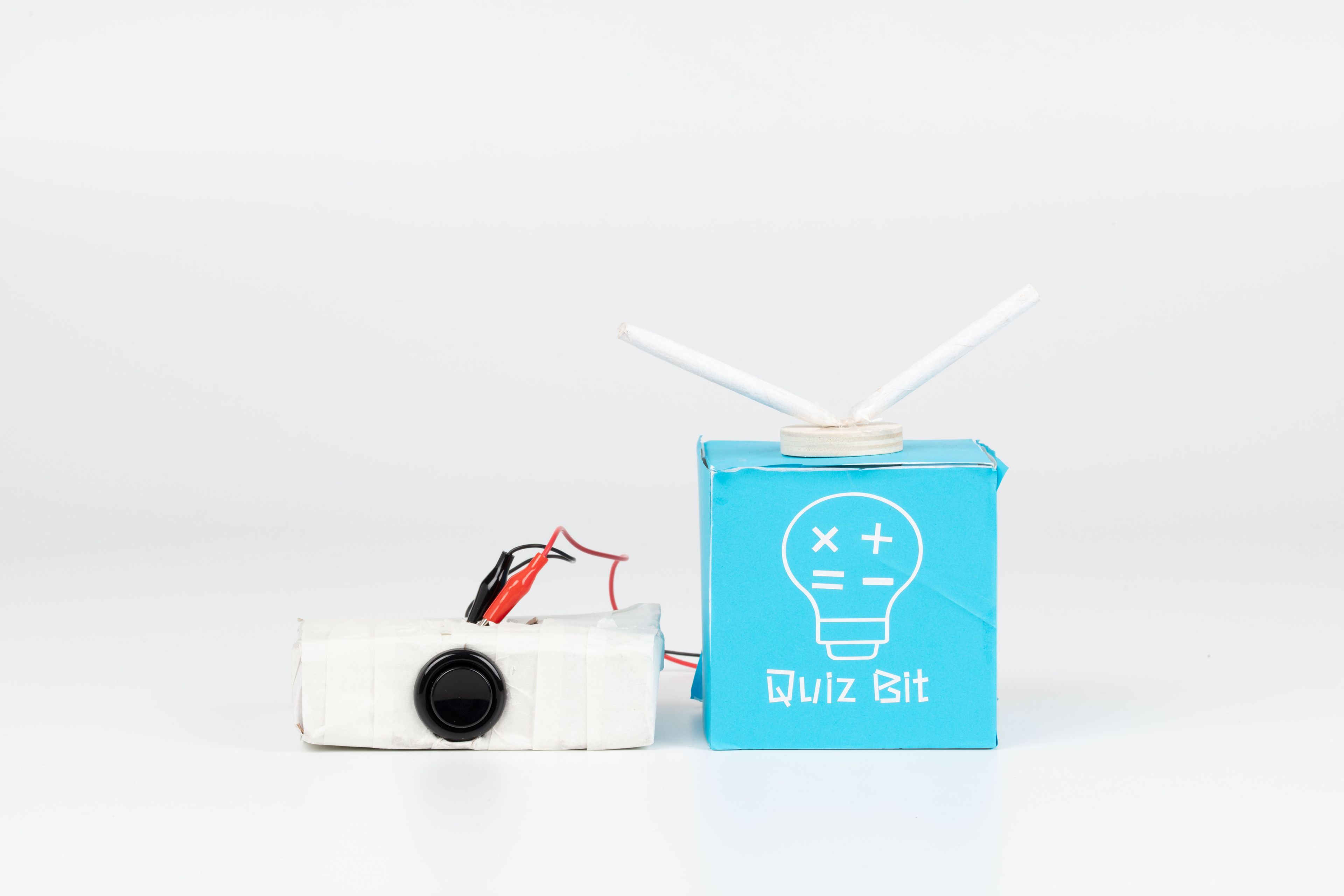 Image of a hand crafted mechanical device against a white background. The device is a blue cube with antennas poking out the top, with red and black wires connected to a taped white box/remote.