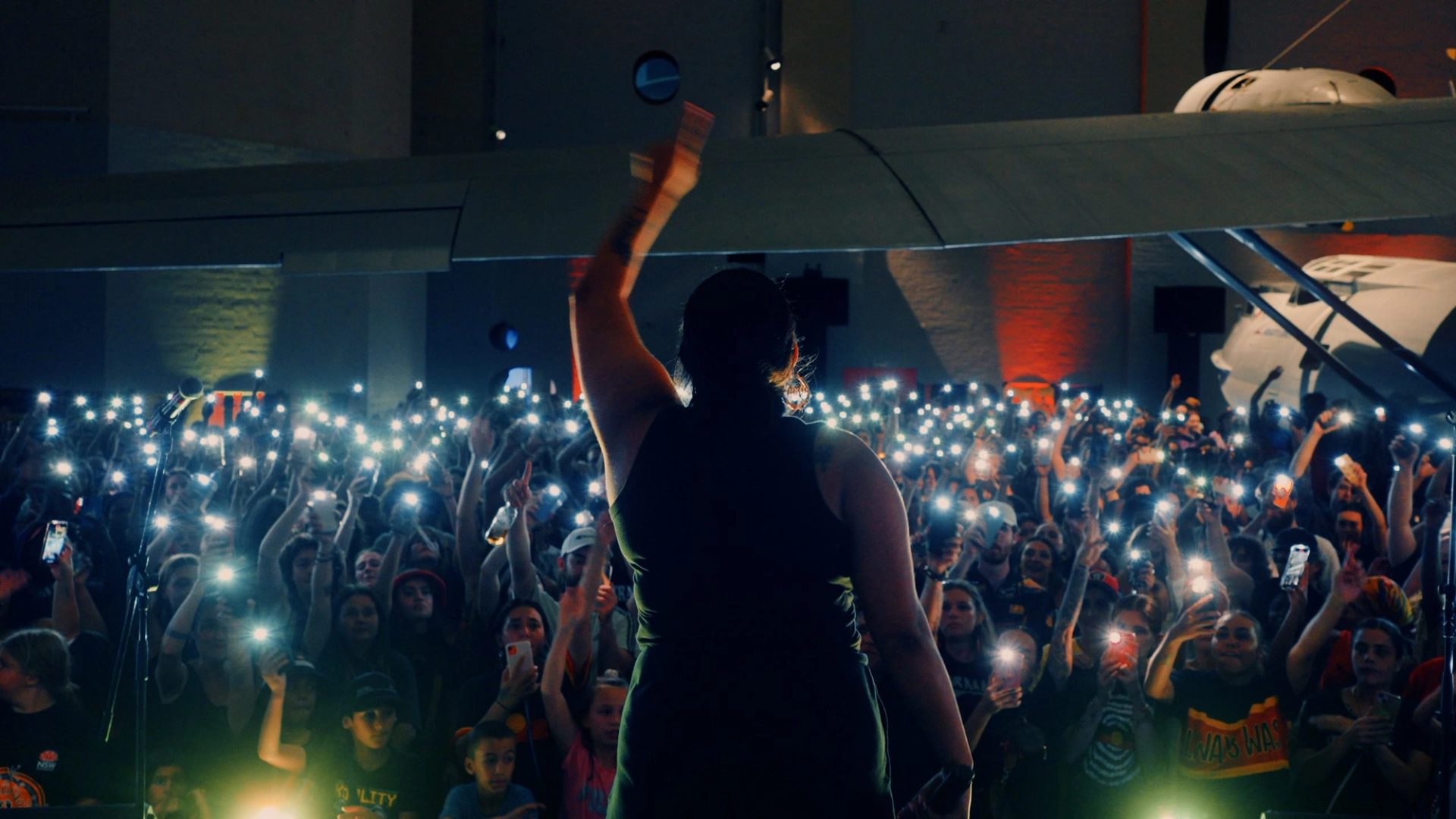 A woman stands on stage in front of a large audience. She has her left hand raised in the air and a microphone in her right hand. The audience are holding their phones up recording the woman.