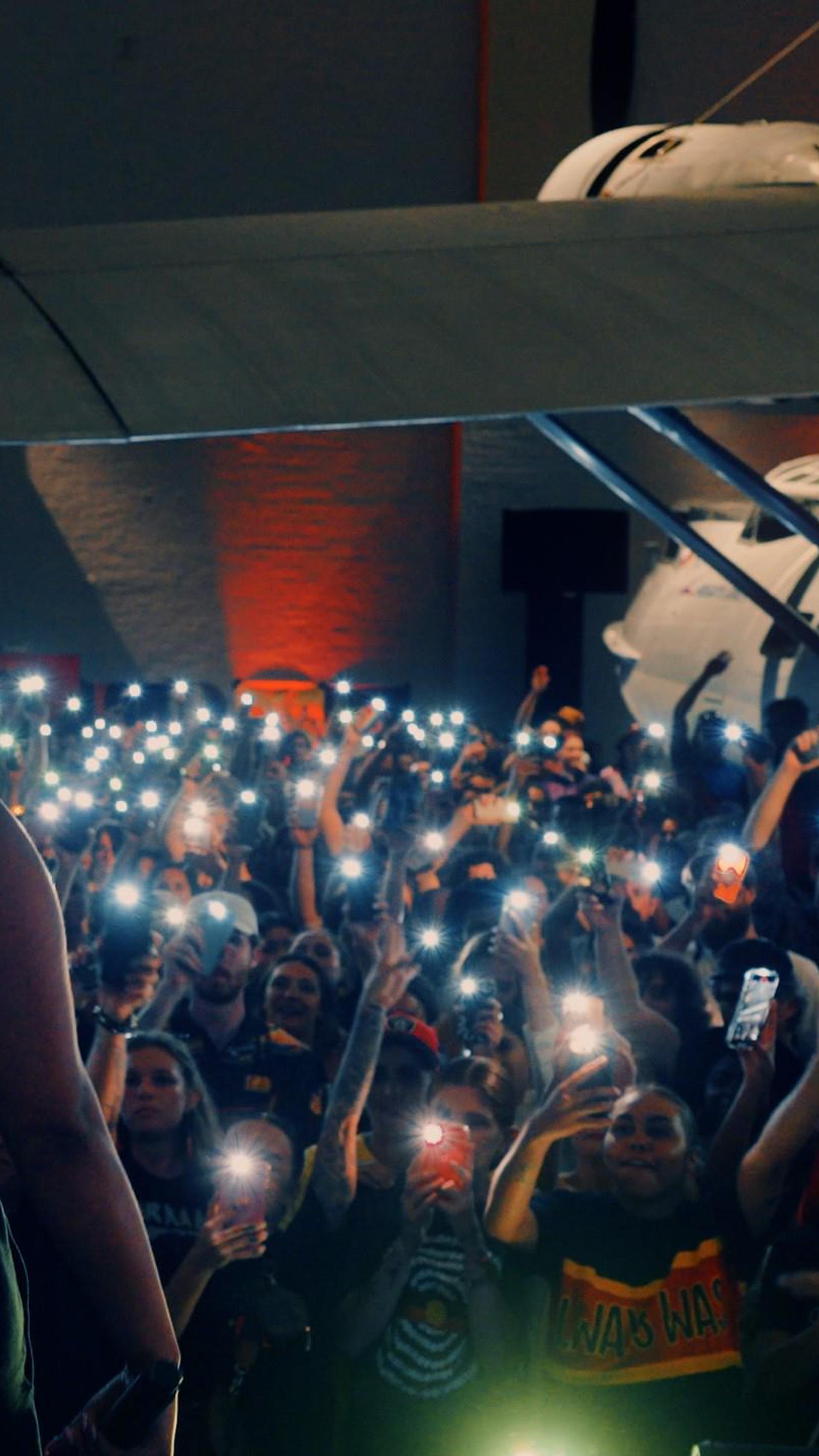 A woman stands on stage in front of a large audience. She has her left hand raised in the air and a microphone in her right hand. The audience are holding their phones up recording the woman.