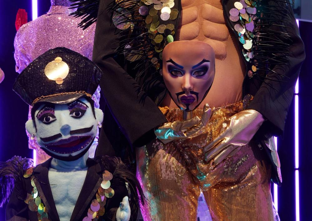 A blue puppet with a black moustache, a black hat with gold trim and a matching open breasted jacket stands next to the bottom half of a mannequin wearing sparkly gold pants and holding a painted face mask.