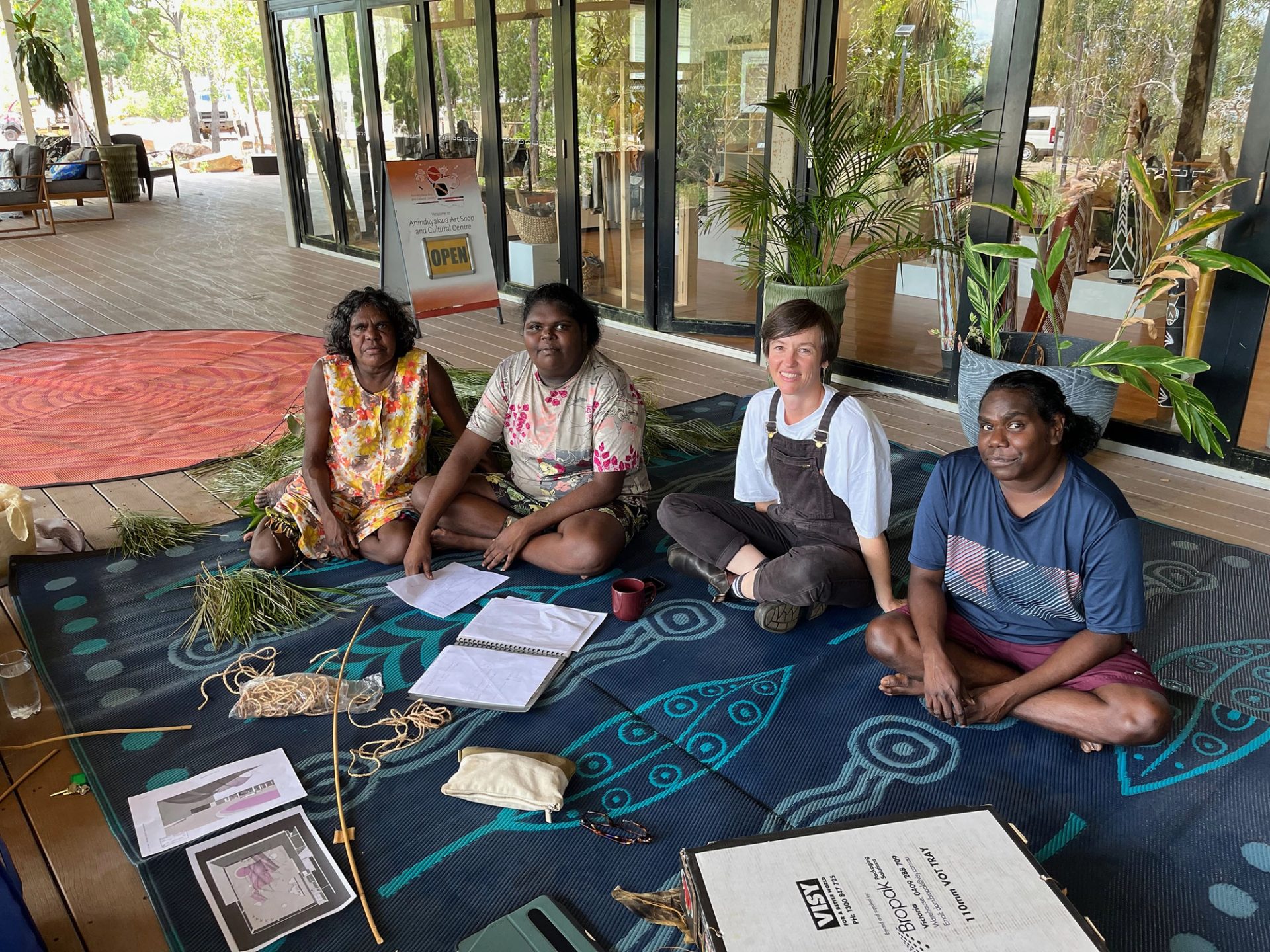 Four women sit on a mat outside an art centre. They are looking at the camera and there is various plant material in front of them.