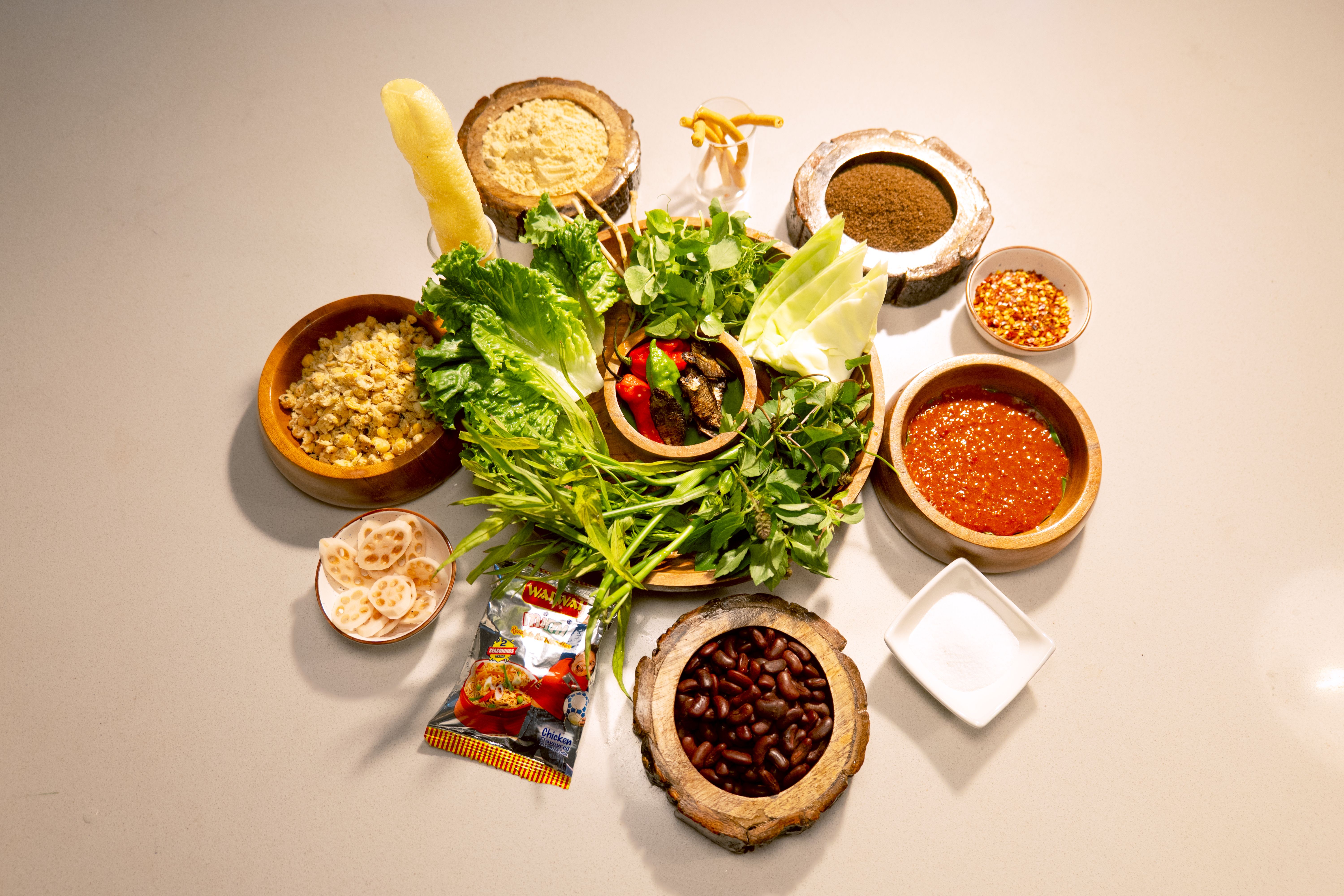 The ingredients for singju. In the centre, a large wooden serving bowl filled with a variety of green vegetables, fresh herbs and chillis. On the outside, smaller wooden and ceramic bowls with kidney beans, dried herbs, spices and condiments.