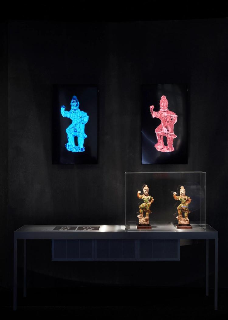 Two small ceramic warrior figures on a silver plinth, above them are blue and red x-ray scans.