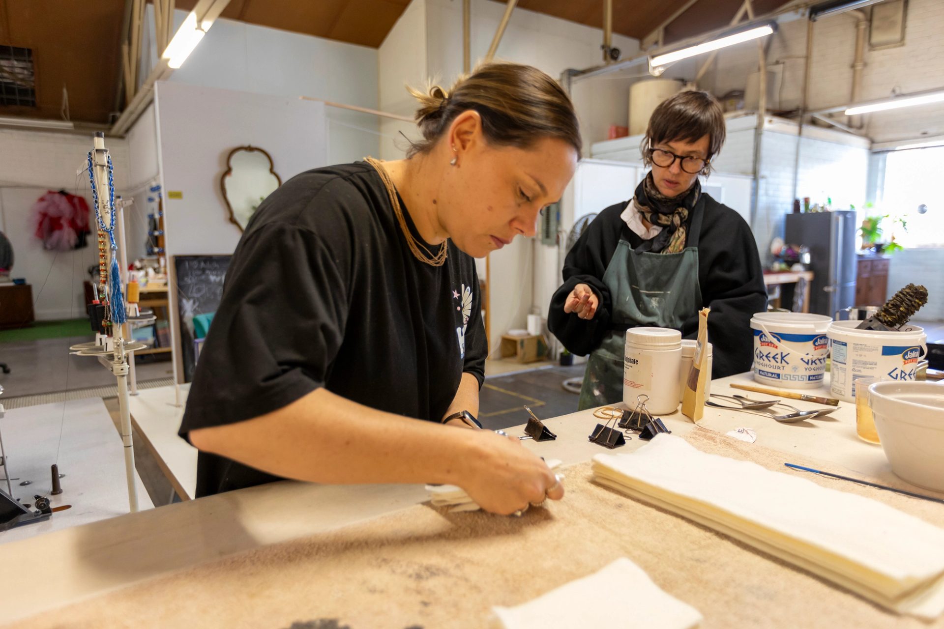 A woman standing over a workshop table, measuring a piece of undyed textile. There is another woman standing behind and to the left of her.