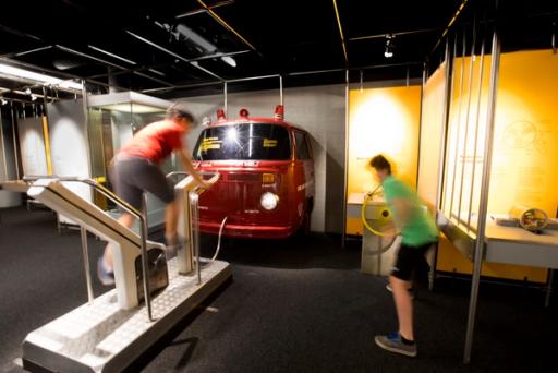 Two children interact with the Experimentations exhibition. Behind them is a front of an old style red fire truck.