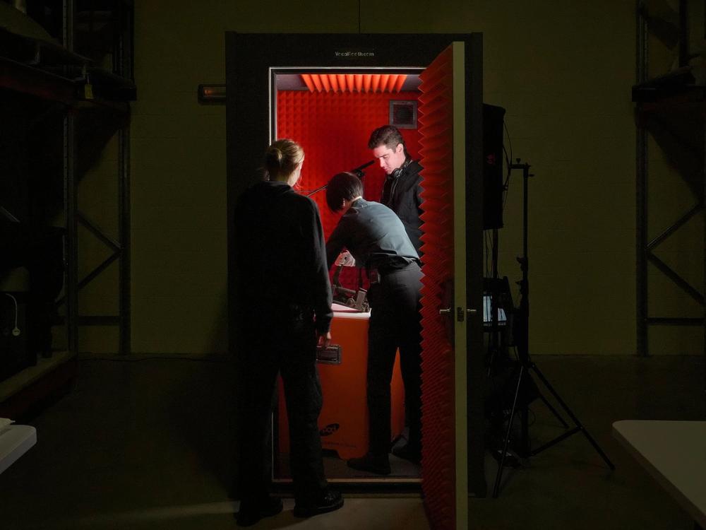 An open door in a dark room allows a glimpse into an orange sound booth. Three figures dressed in black stand in and near the doorway, huddled around an object.