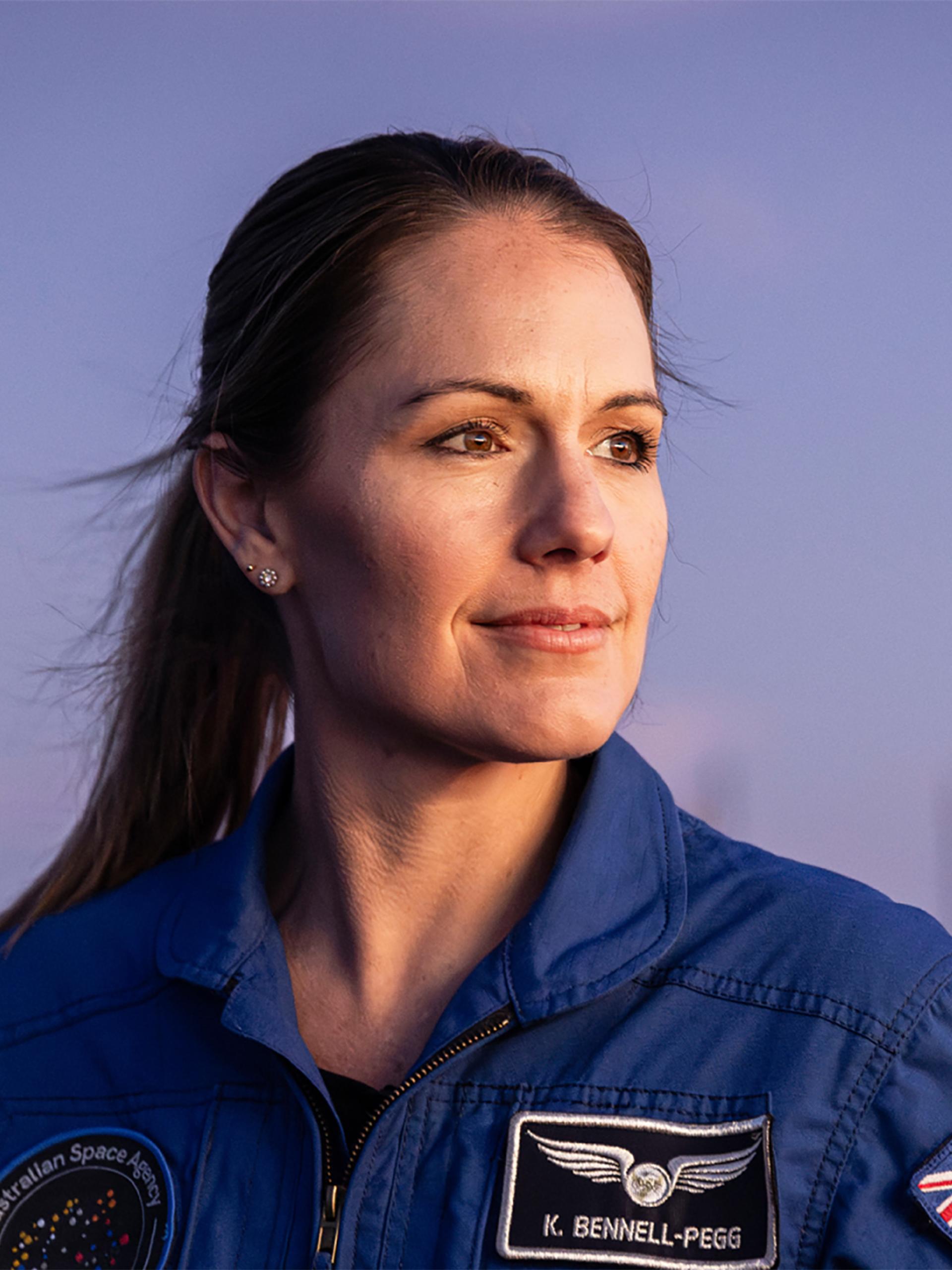 Astronaut Katherine Bennell-Pegg posed against a blue sky, looking to her left in a blue space jumpsuit.