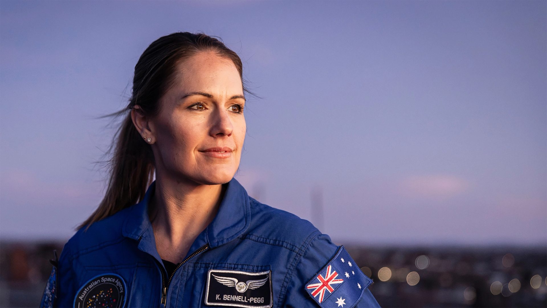 Astronaut Katherine Bennell-Pegg posed against a blue sky, looking to her left in a blue space jumpsuit.