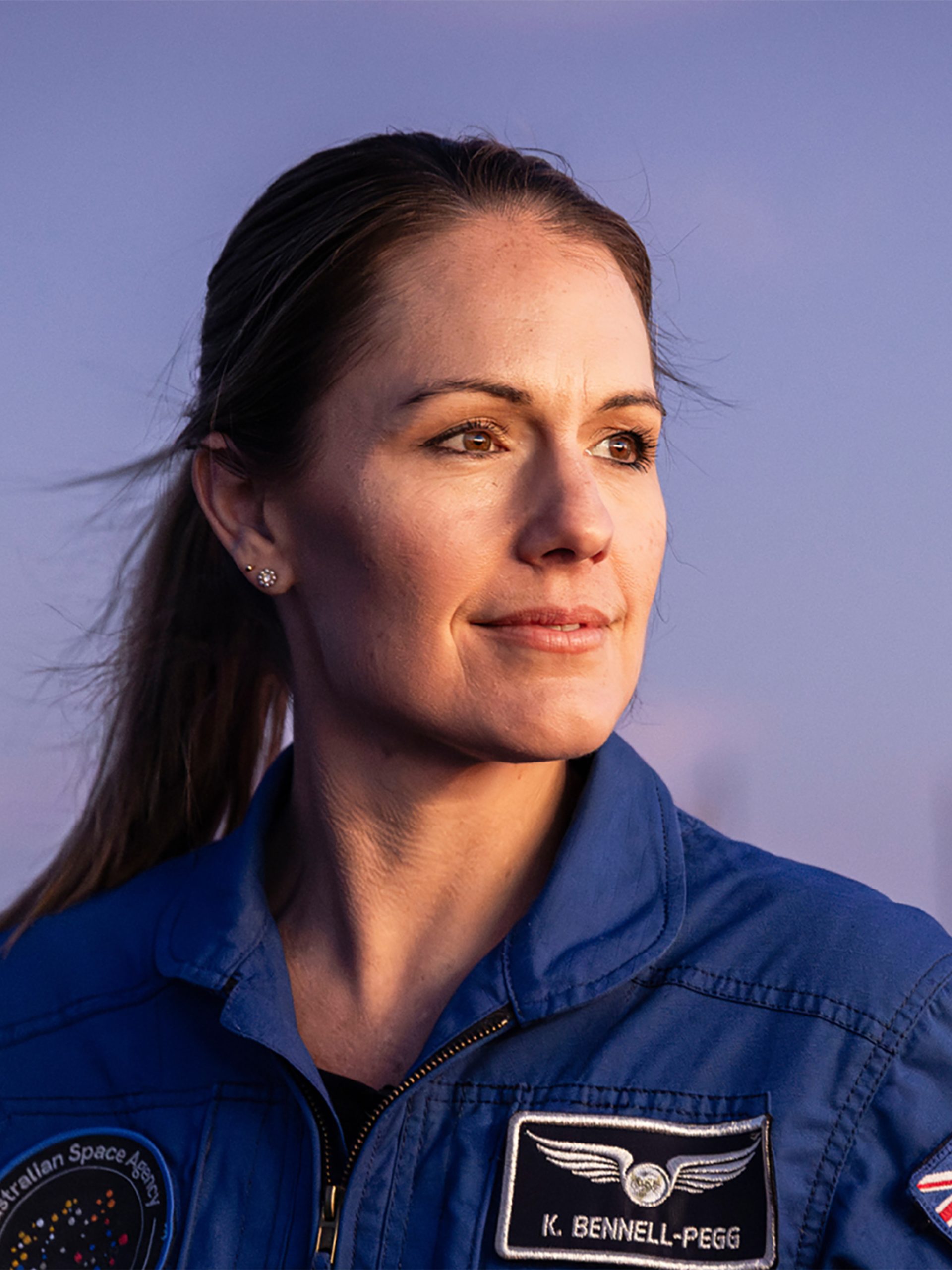 Astronaut Katherine Bennell-Pegg posed against a blue sky, looking to her left in a blue space jumpsuit.