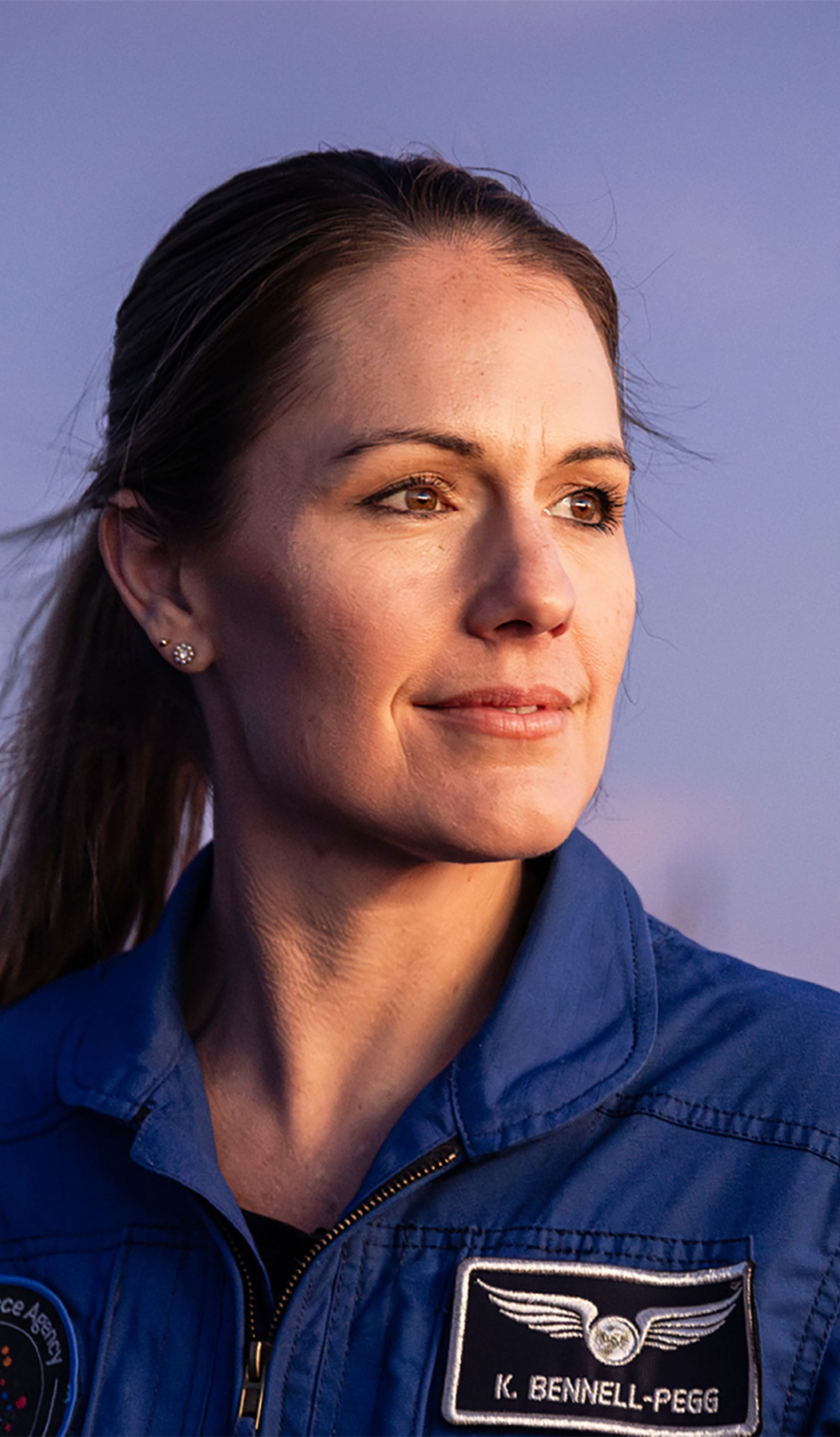 Astronaut Katherine Bennell-Pegg posed against a blue sky, looking to her left in a blue space jumpsuit.