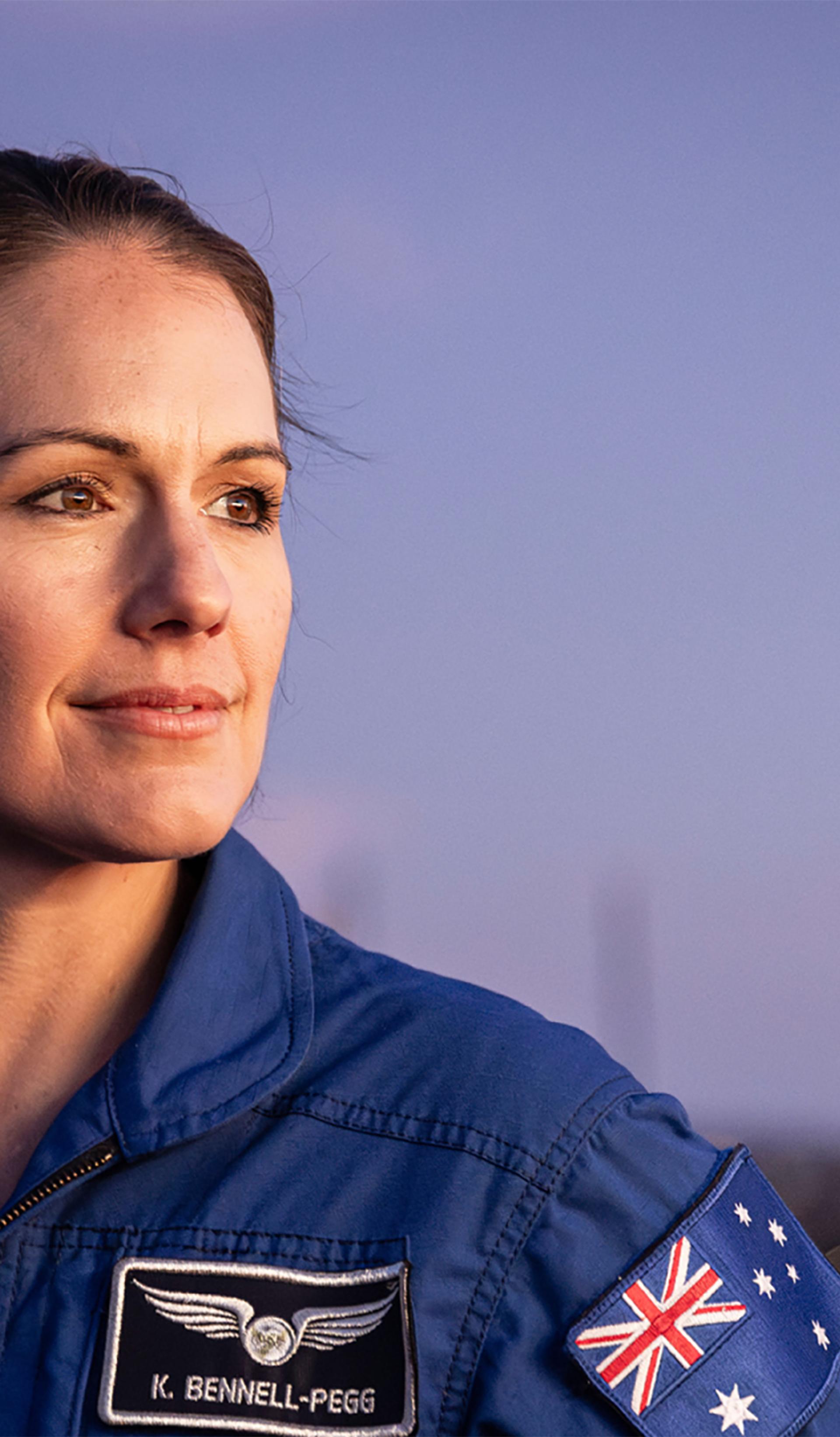 Astronaut Katherine Bennell-Pegg posed against a blue sky, looking to her left in a blue space jumpsuit.
