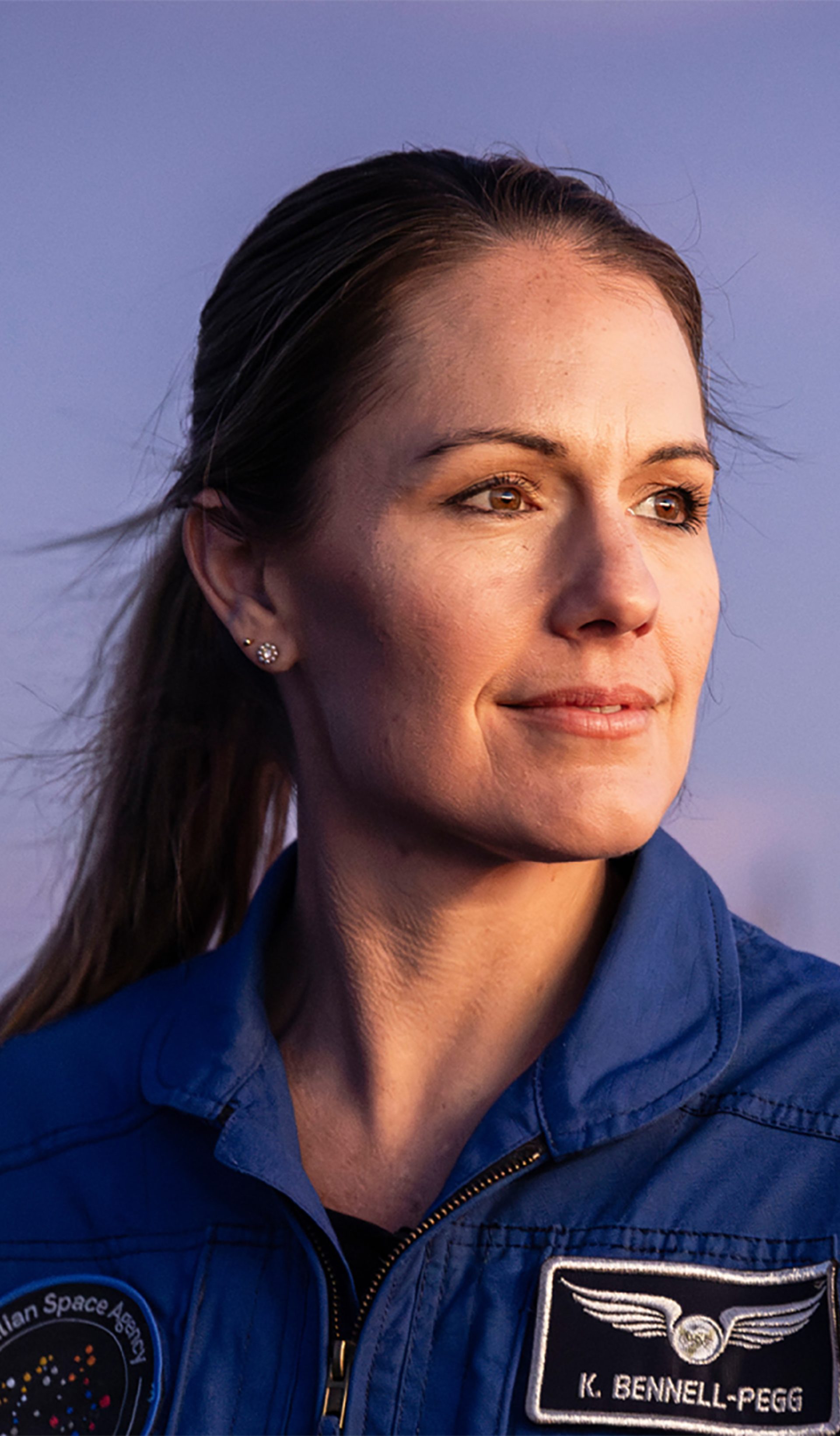 Astronaut Katherine Bennell-Pegg posed against a blue sky, looking to her left in a blue space jumpsuit.