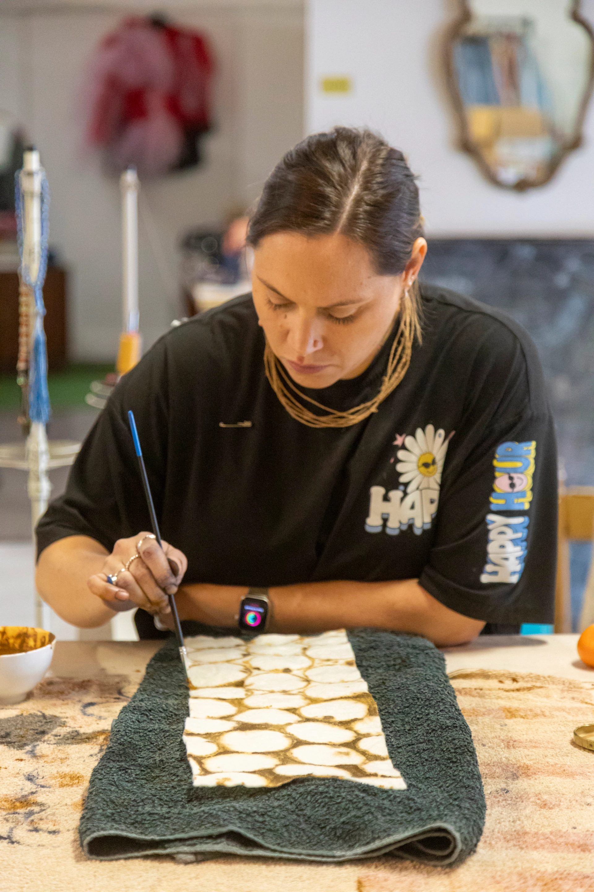 A woman leans over a work bench and is using a thin paintbrush to paint a circular pattern on to a piece of textile.