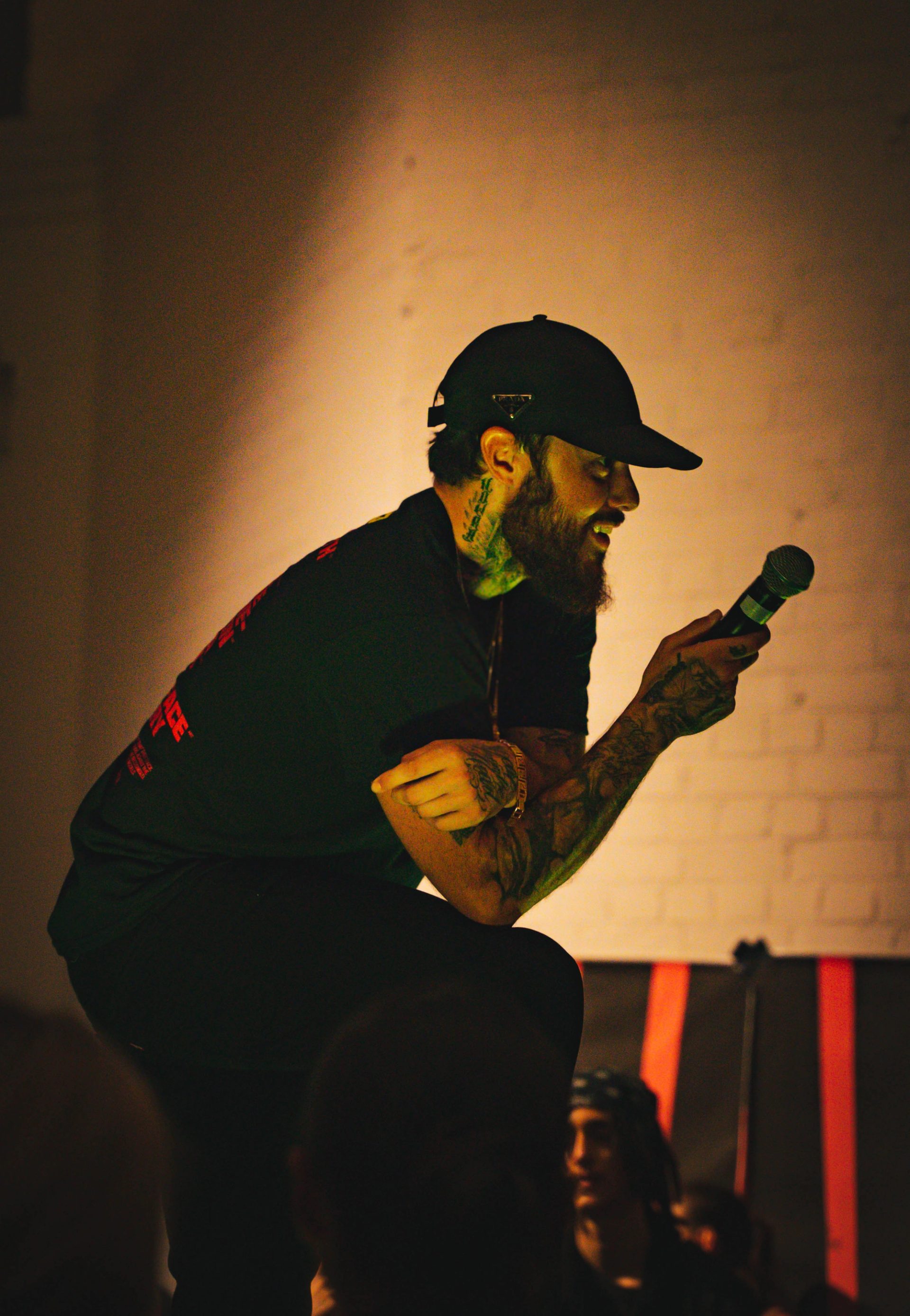 A man leaning forward and resting his right arm on his right knee. He is on stage for a performance and is holding a microphone in his right hand.