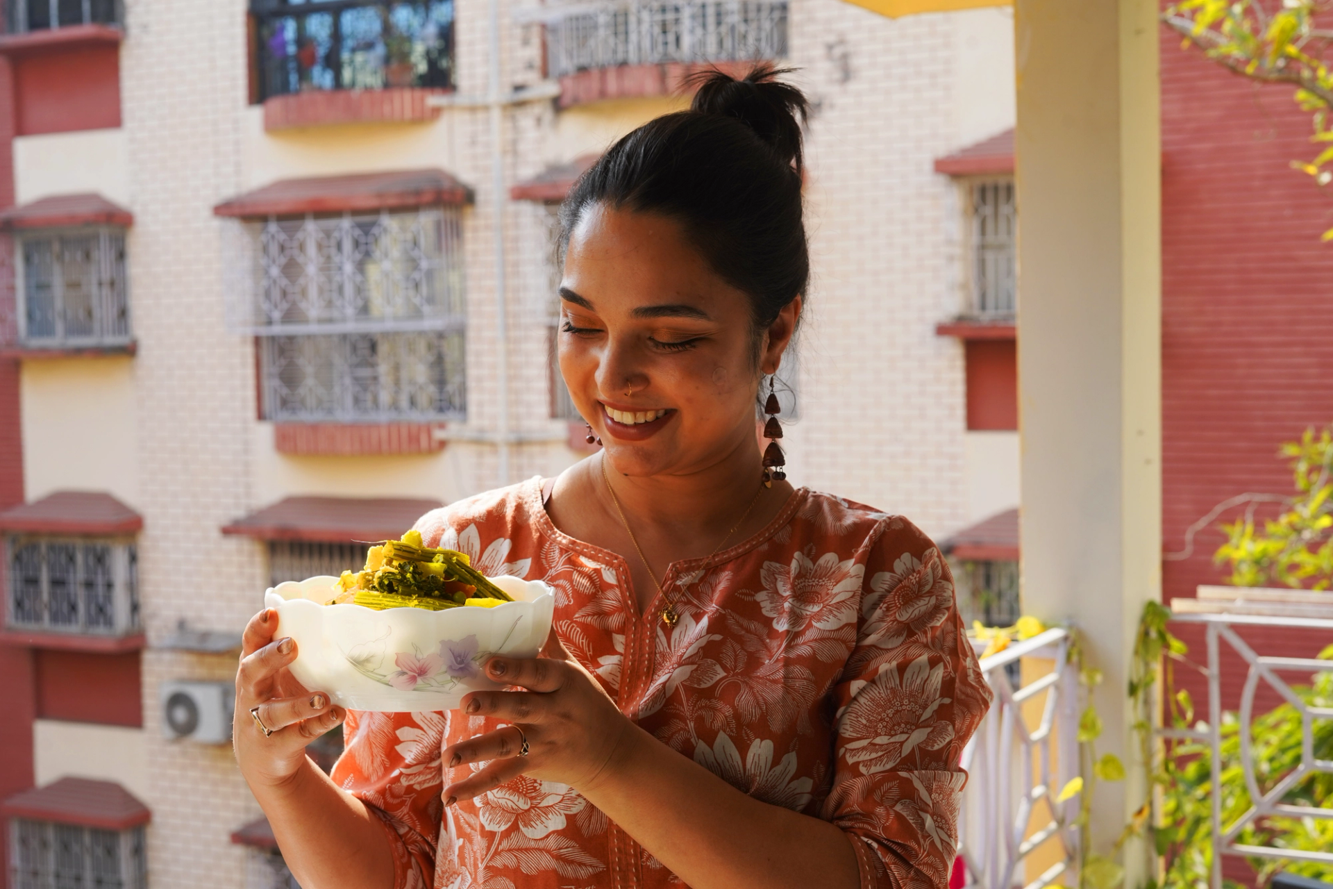 A Bengali woman stands on her apartment balcony and holds a bowl of vegetable stir-fry. She wears a pink top with white floral print and drop earrings. She is looking at the stir-fry and smiling.