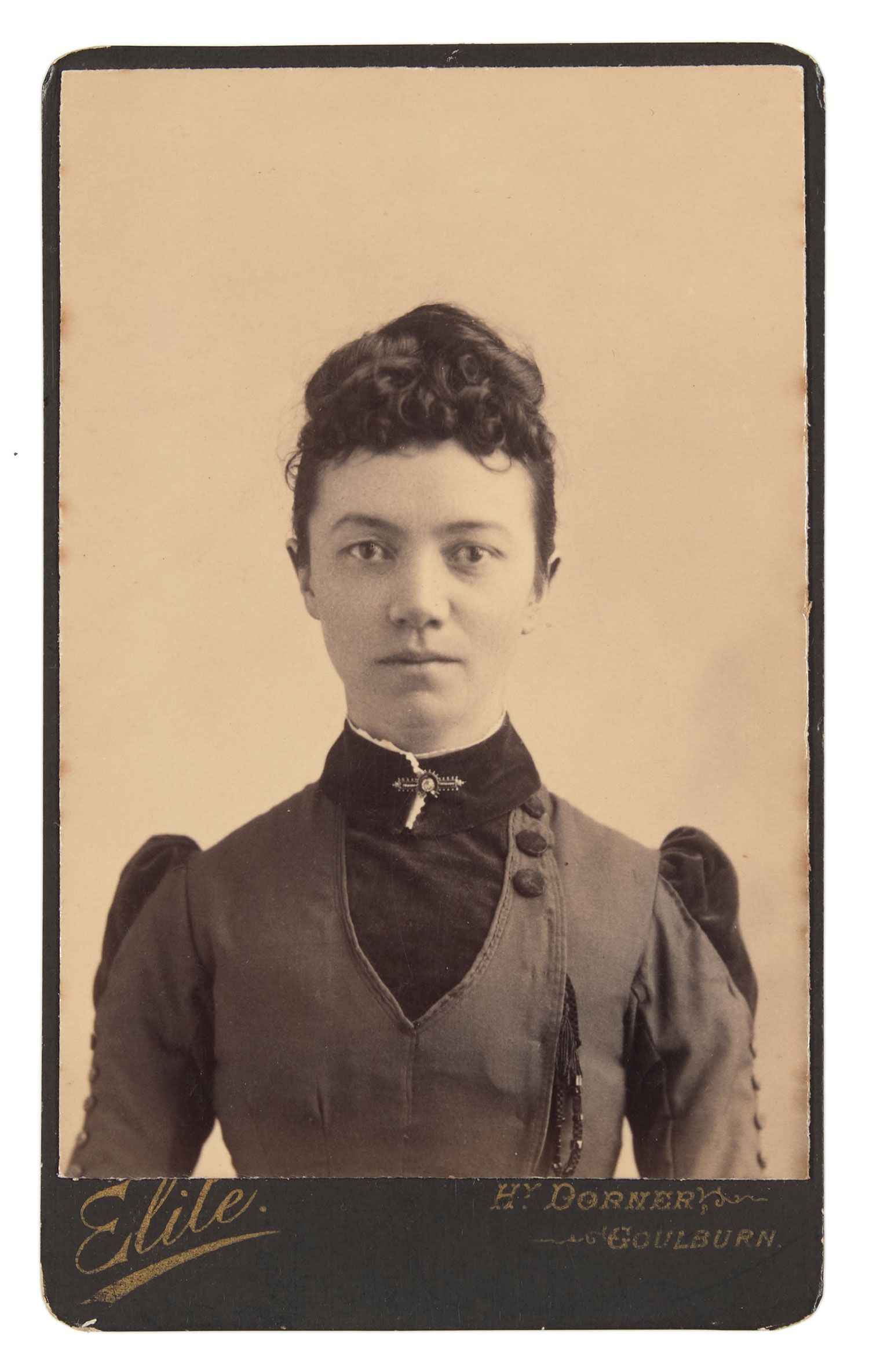 Mounted black and white photograph depicting a girl facing straight to the camera. She is wearing a high neck, long sleeved dress with puffs on the shoulders.