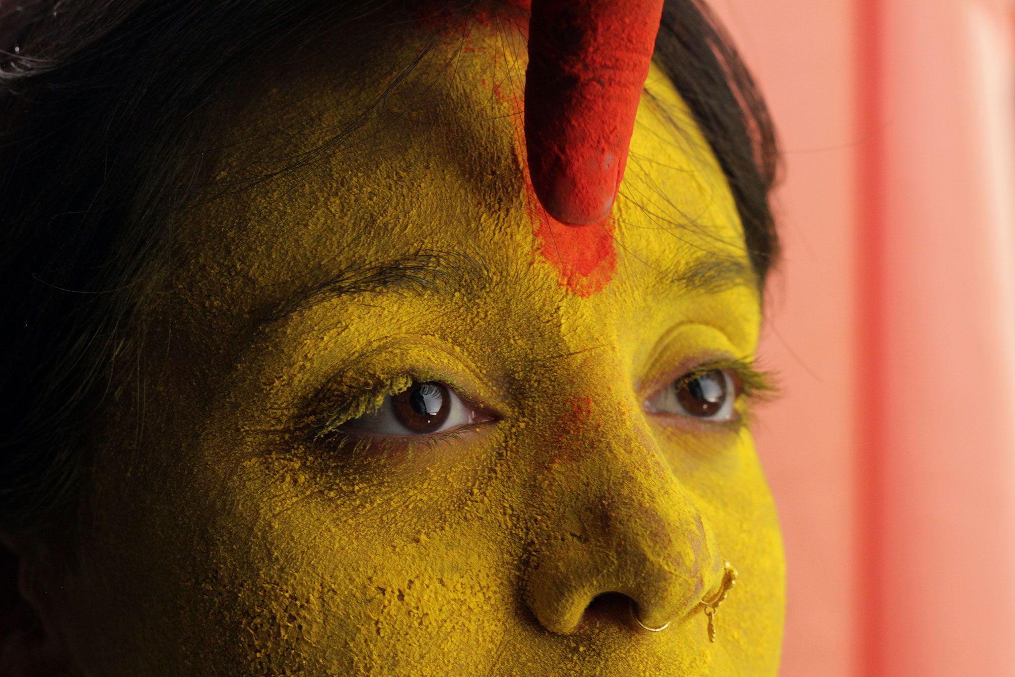 A close up of a woman’s face covered in yellow pigment. A finger is marking a red line on her forehead.