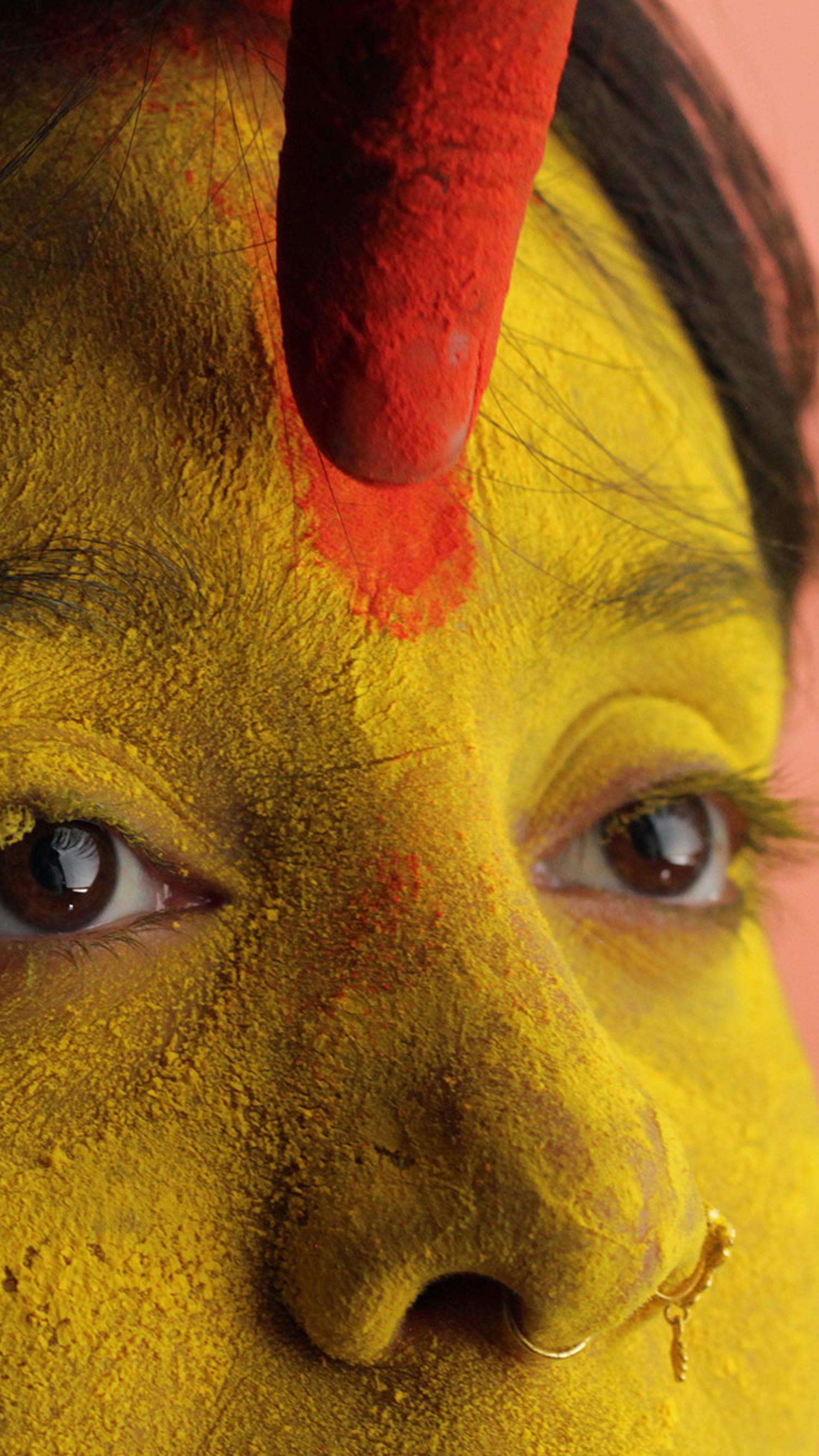 A close up of a woman’s face covered in yellow pigment. A finger is marking a red line on her forehead.