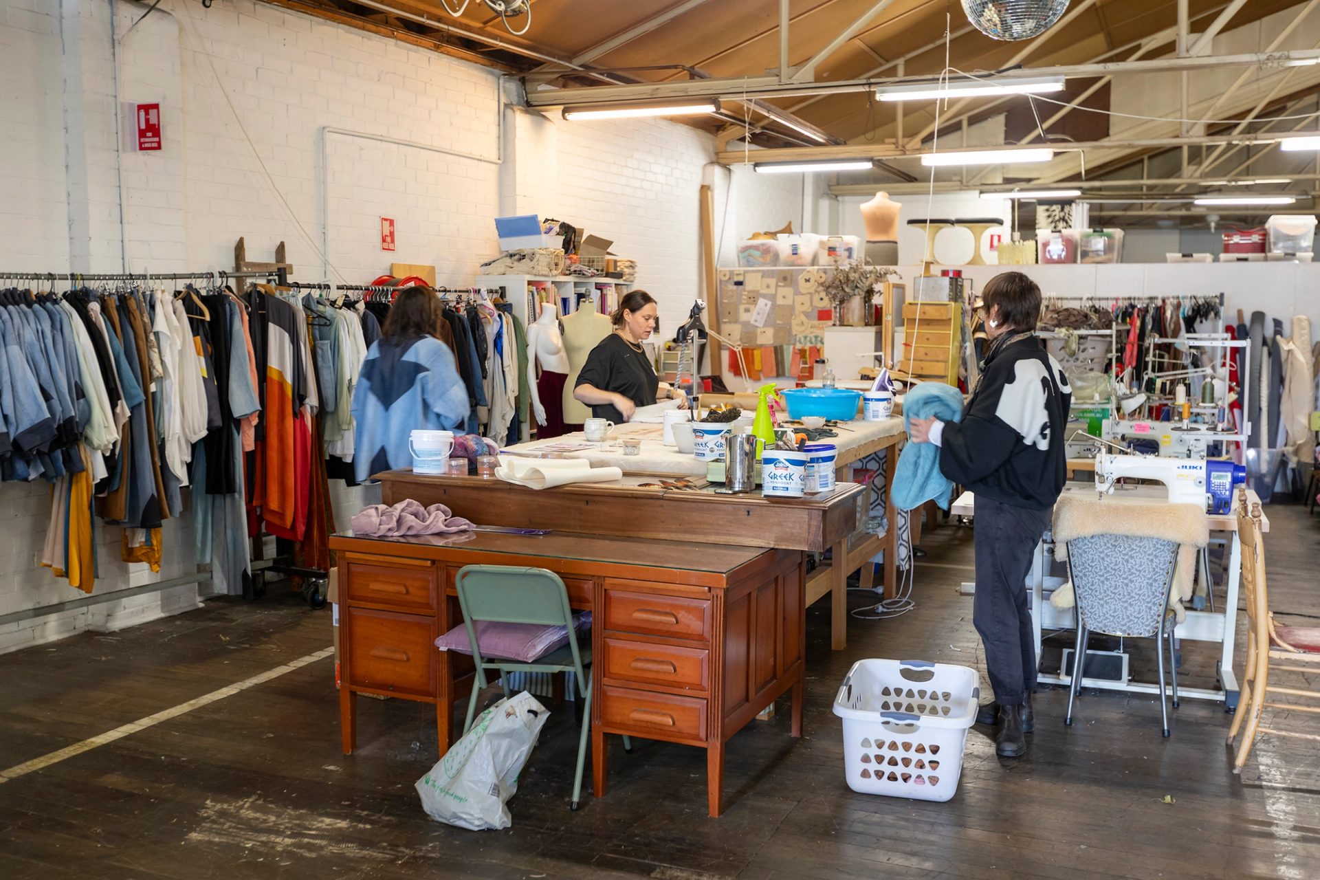 A large studio with racks of clothes lining the walls and three women standing around a large table covered with various fabrics and textile tools.