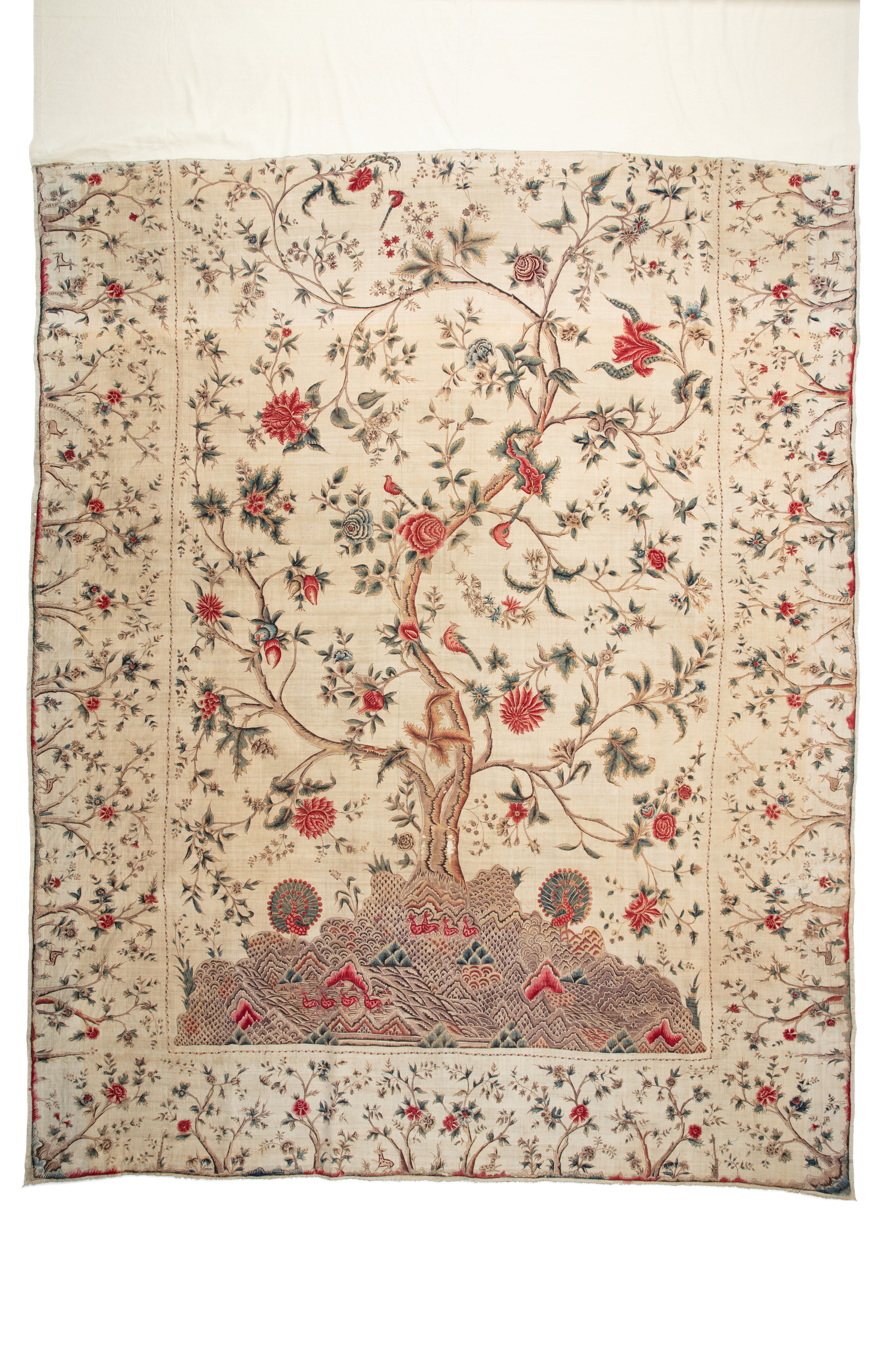 Bed hanging made of cream-coloured hand spun and hand woven cotton calico. The design is of the Tree of Life and has been hand painted using kalamkari penwork and plant dyeing with mordants. The design depicts a large central tree populated with birds, trailing flowers and leaves. The foothills below the tree show intricate details of deer, scenery and two peacocks. The tree borders depict small trees, flowers and more rambling deer.