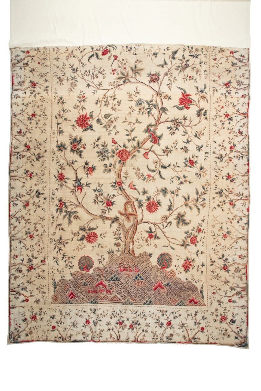 Bed hanging made of cream-coloured hand spun and hand woven cotton calico. The design is of the Tree of Life and has been hand painted using kalamkari penwork and plant dyeing with mordants. The design depicts a large central tree populated with birds, trailing flowers and leaves. The foothills below the tree show intricate details of deer, scenery and two peacocks. The tree borders depict small trees, flowers and more rambling deer.