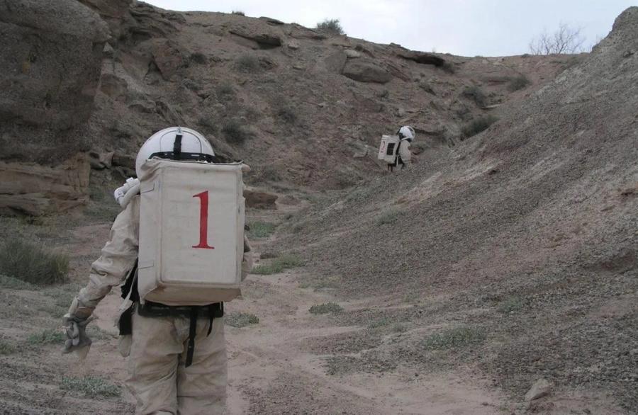 Two figures wearing space suits walk amongst a desert ranch.