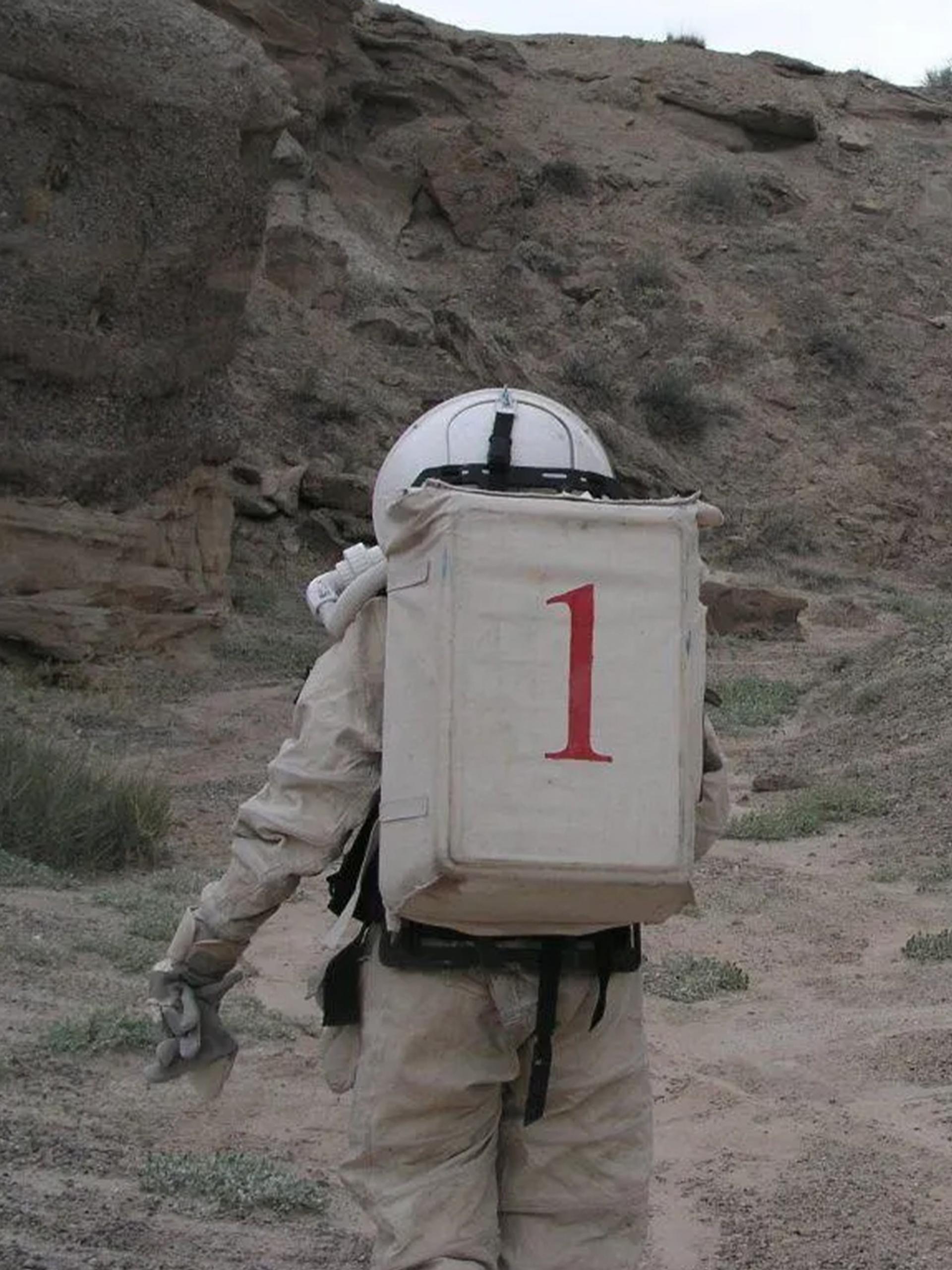 Two figures wearing space suits walk amongst a desert ranch.