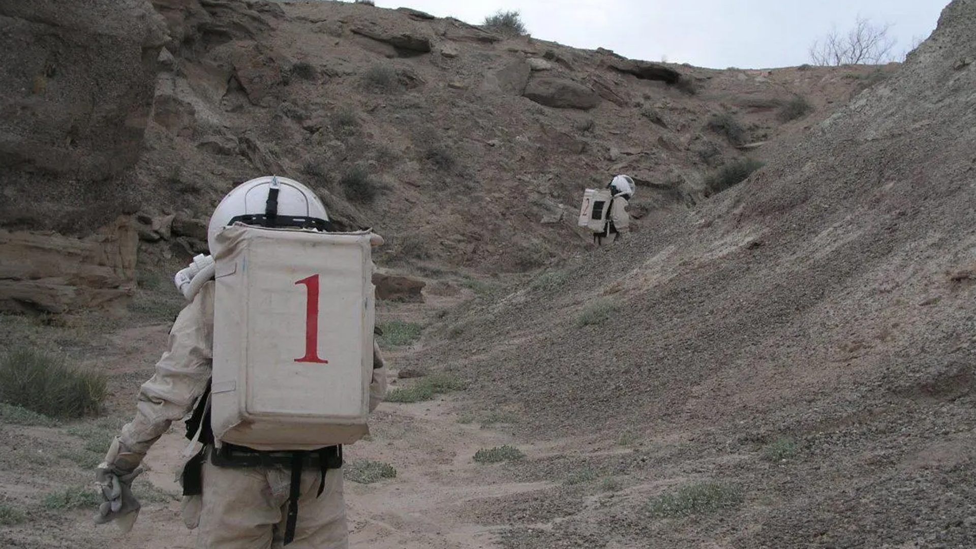 Two figures wearing space suits walk amongst a desert ranch.