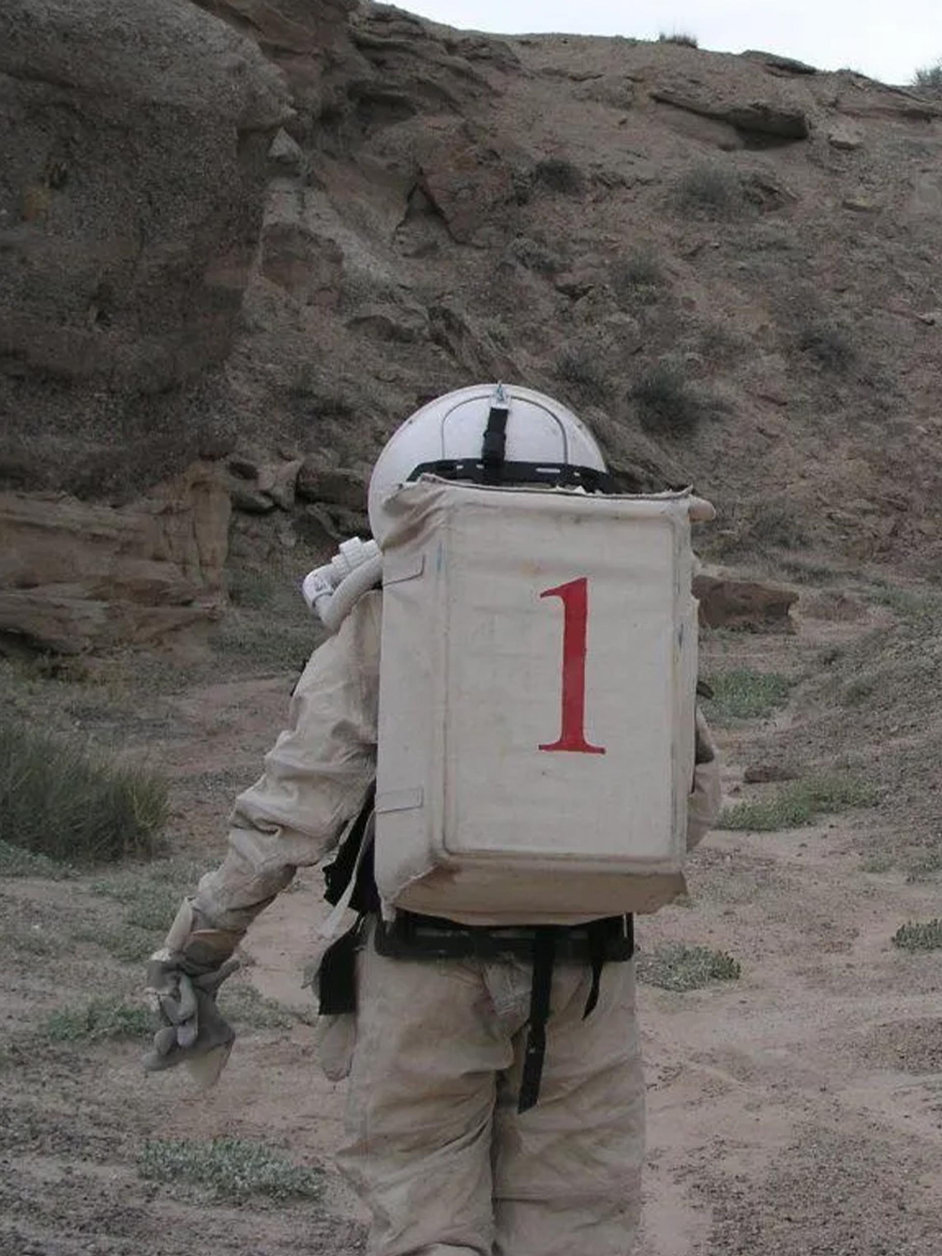 Two figures wearing space suits walk amongst a desert ranch.
