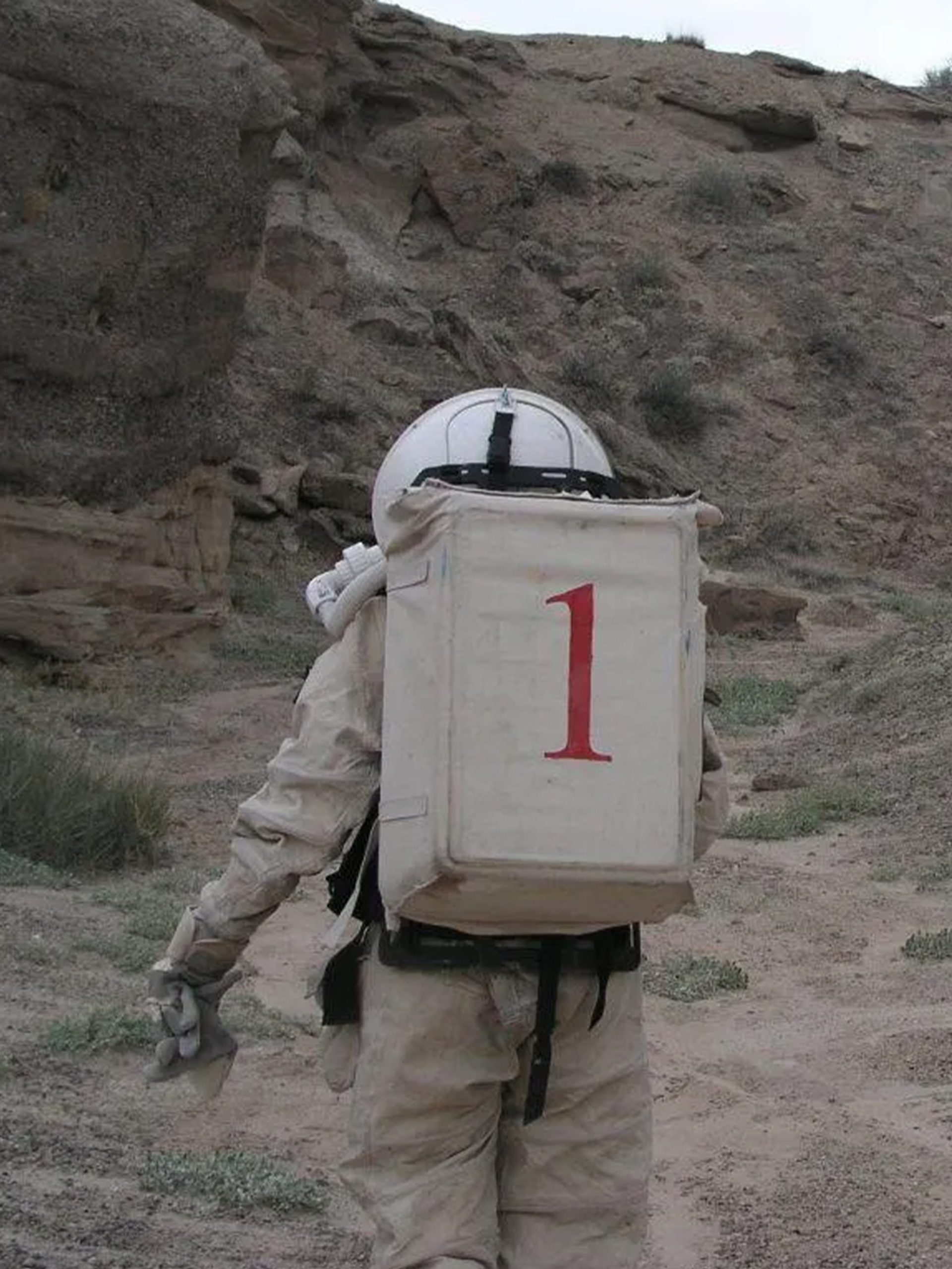 Two figures wearing space suits walk amongst a desert ranch.