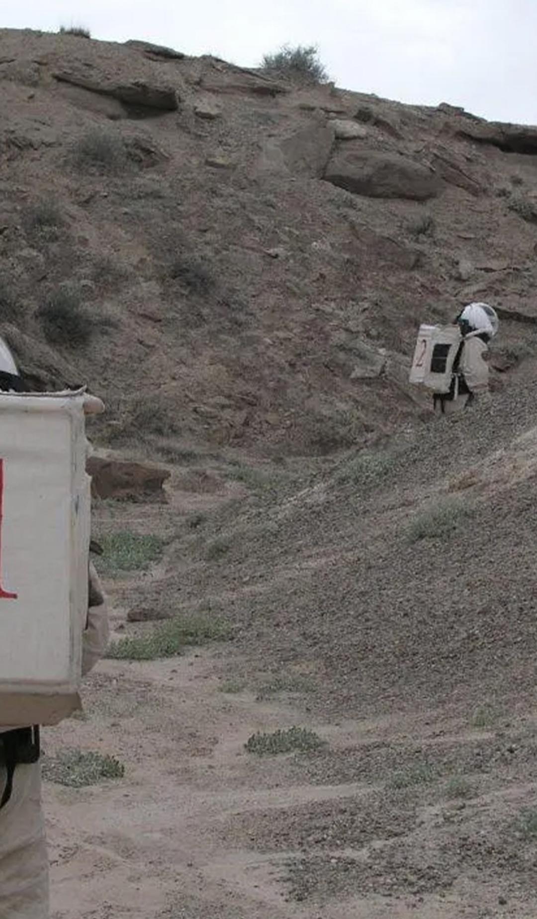Two figures wearing space suits walk amongst a desert ranch.