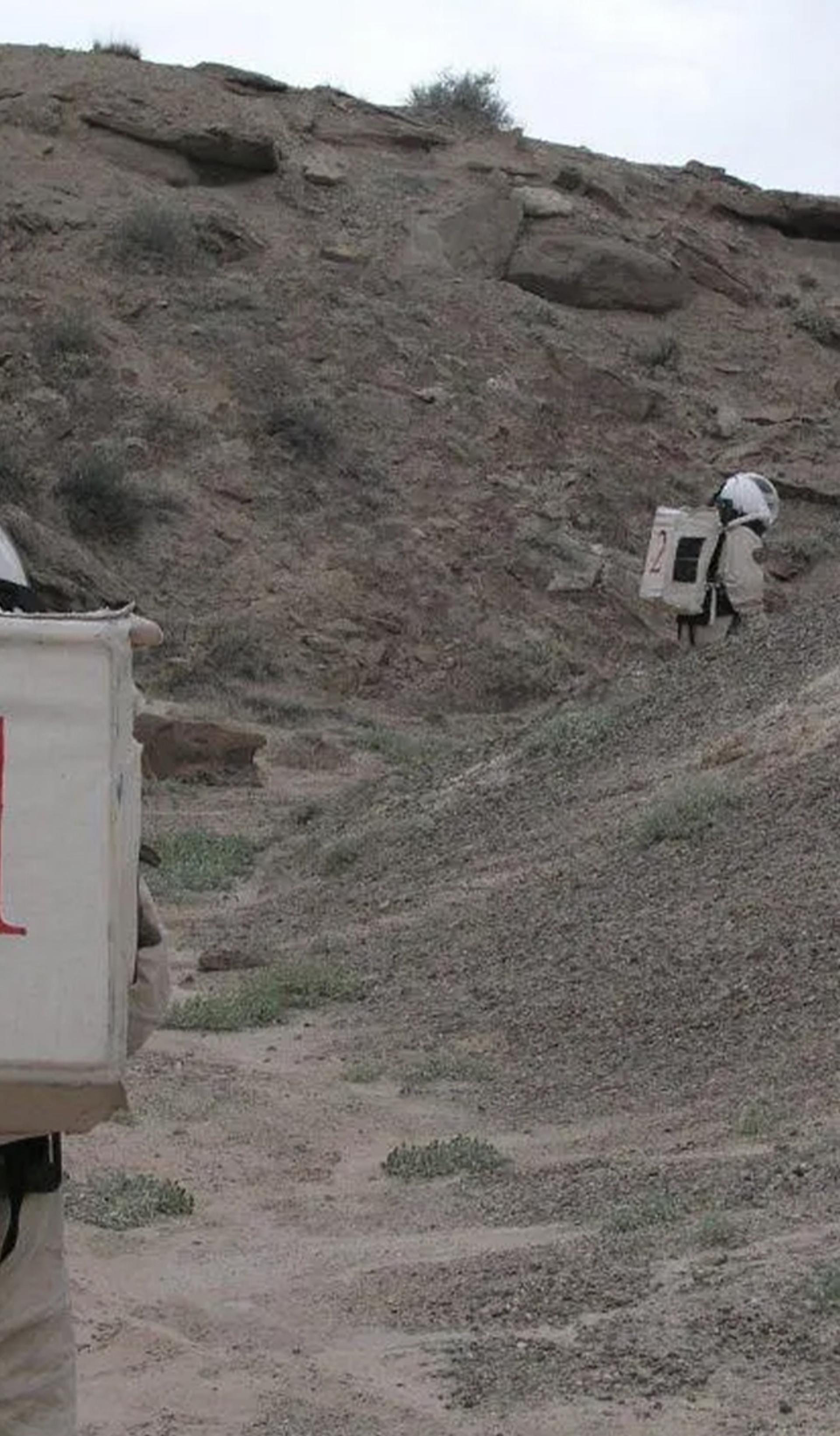 Two figures wearing space suits walk amongst a desert ranch.