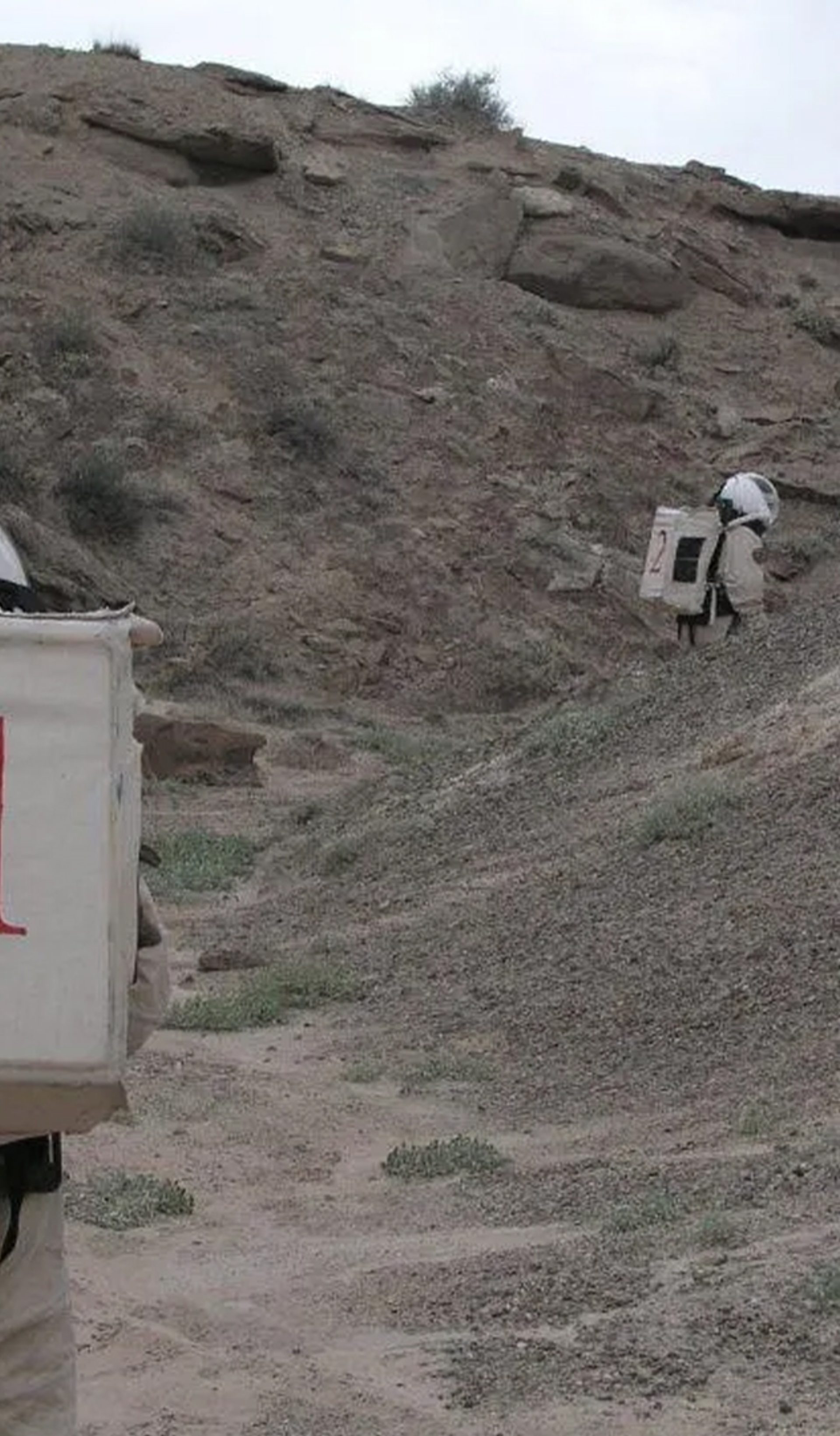 Two figures wearing space suits walk amongst a desert ranch.