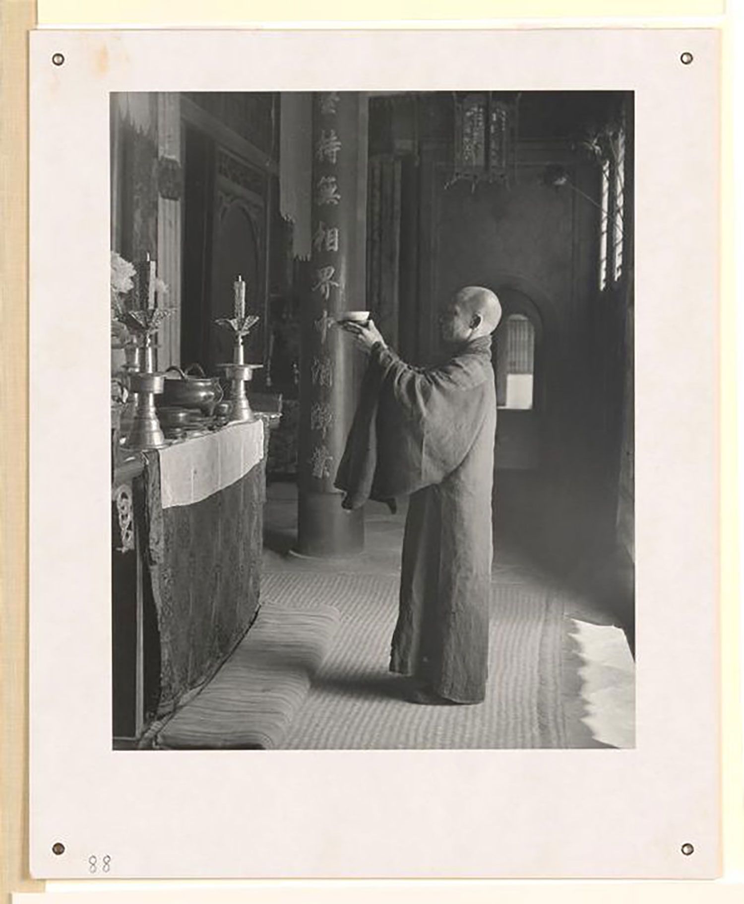 Black and white photograph showing a monk making an offering at an altar in a Buddhist temple. Dressed in a long robe, with a shaven head, he is holding up a bowl in front of the altar. On the altar are two large candle holders and various metal vessels. The altar is situated in a large hall with columns. Couplets carved into the wood adorn the columns.