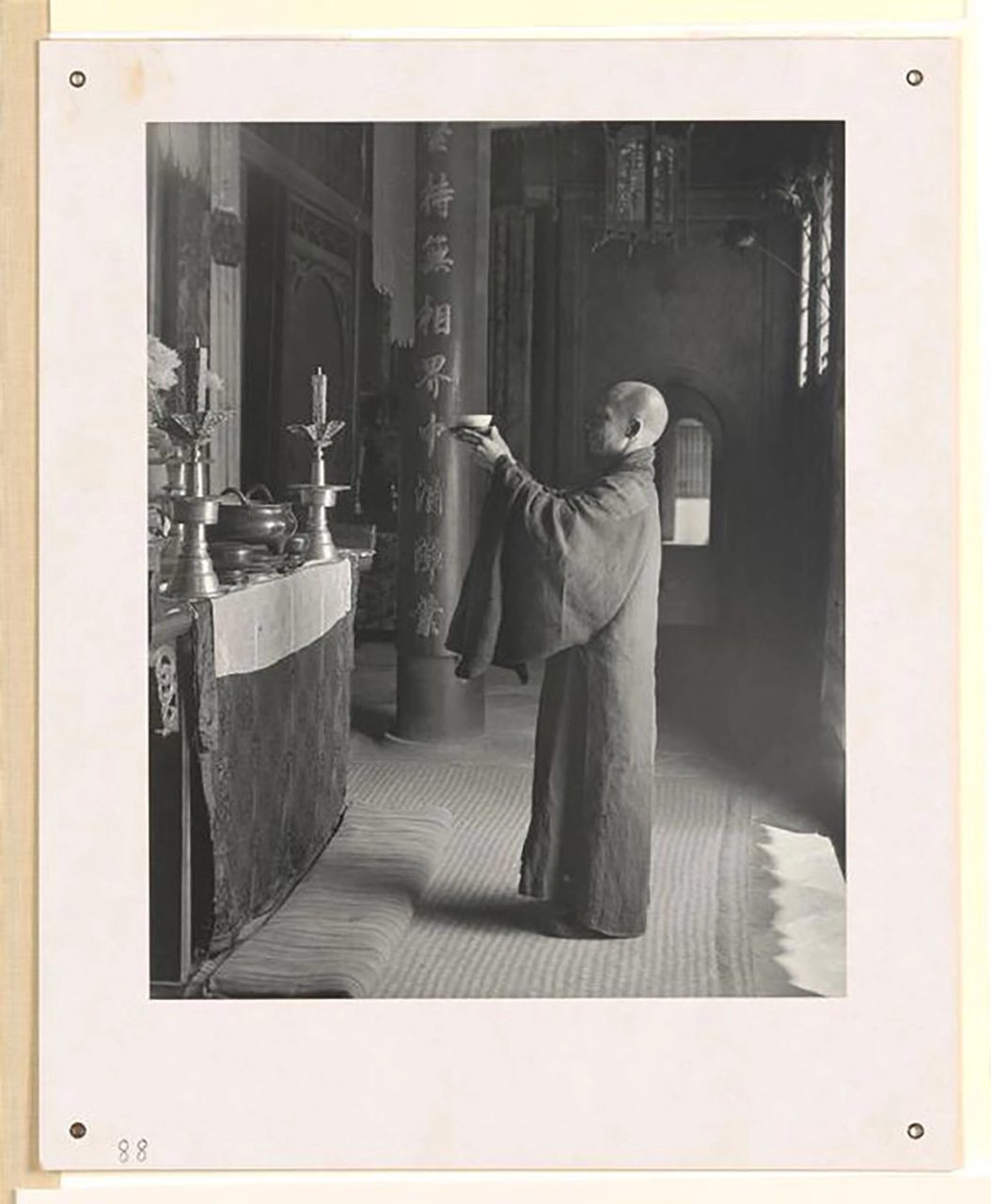 Black and white photograph showing a monk making an offering at an altar in a Buddhist temple. Dressed in a long robe, with a shaven head, he is holding up a bowl in front of the altar. On the altar are two large candle holders and various metal vessels. The altar is situated in a large hall with columns. Couplets carved into the wood adorn the columns.