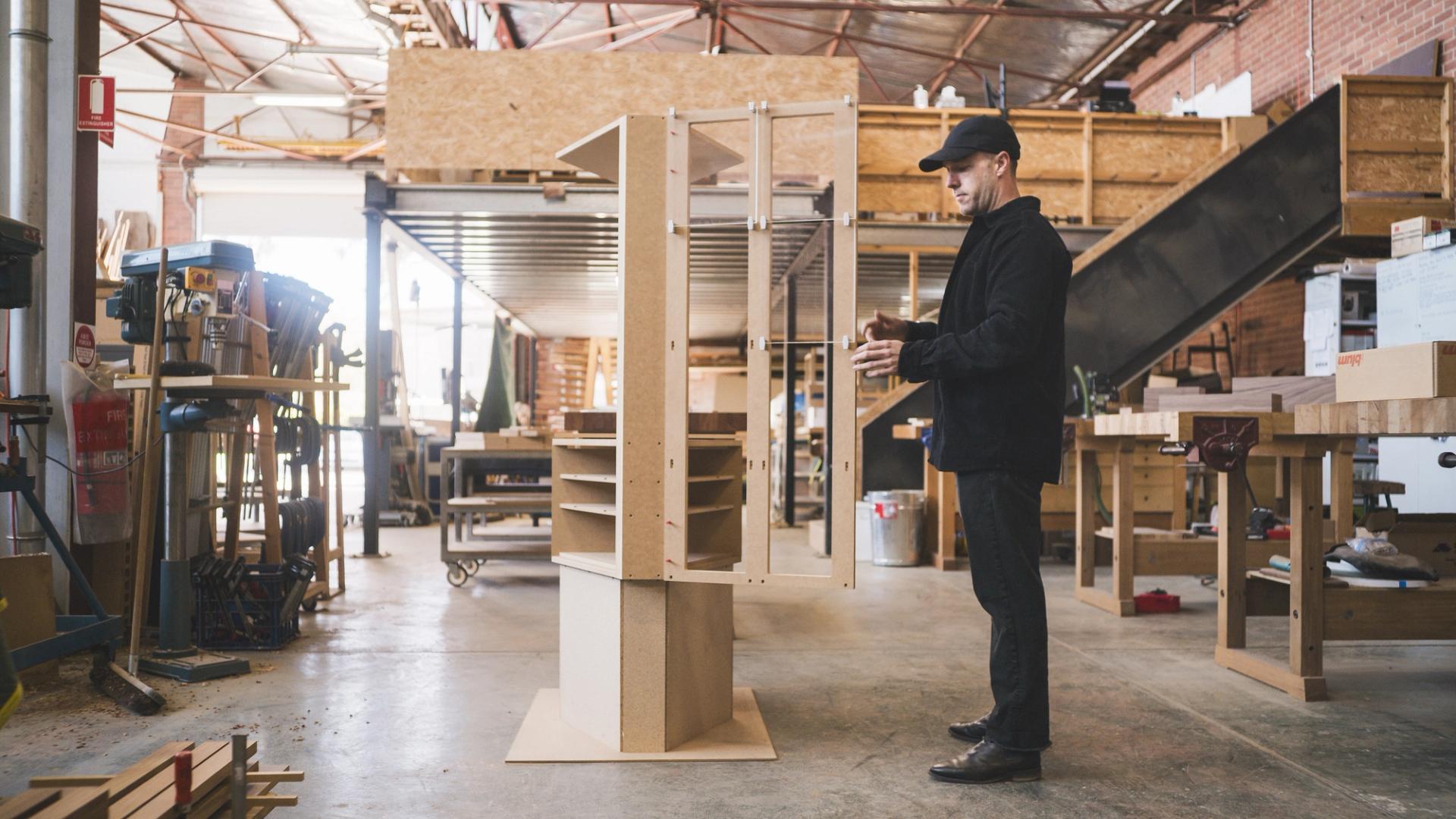In a light-filled furniture workshop, a man interacts with a prototype of a large cabinet. He’s dressed in black.