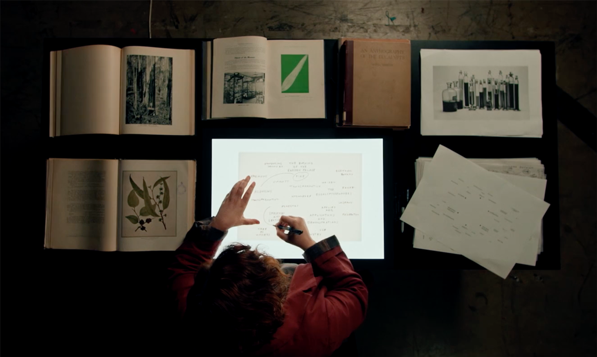 Figure sits at a desk writing on an illuminated surface, at a birds eye view