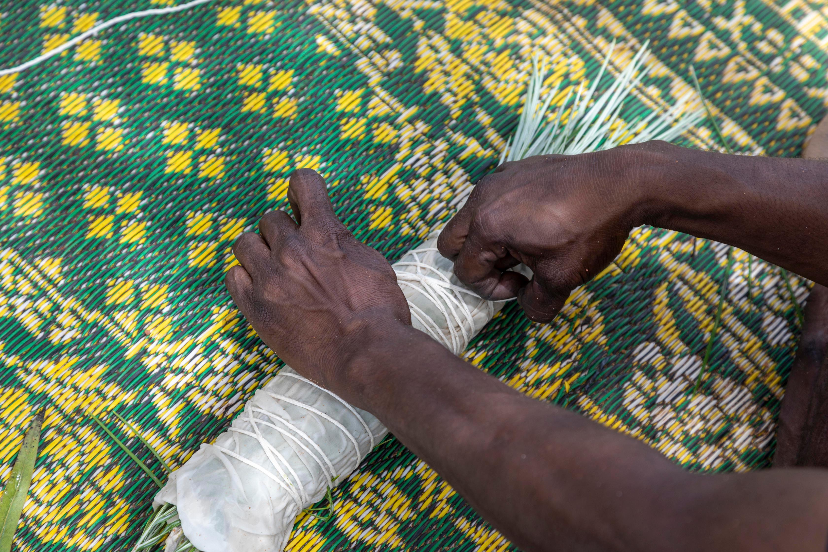 Two hands wrapping a bundle of grass with white fabric and string on a green and yellow mat.