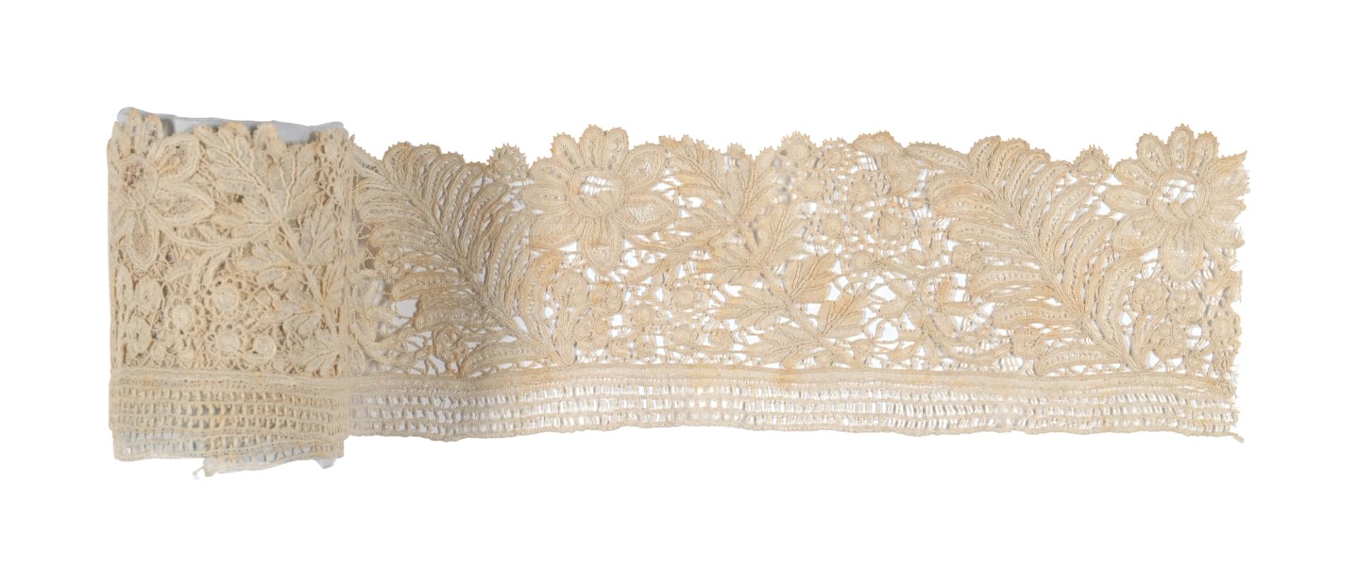 A roll of lace border, bobbin applique on machine made net with needle lace filling, made of cotton. Depicts a scrolling floral design with a 3 row square grid edge.
